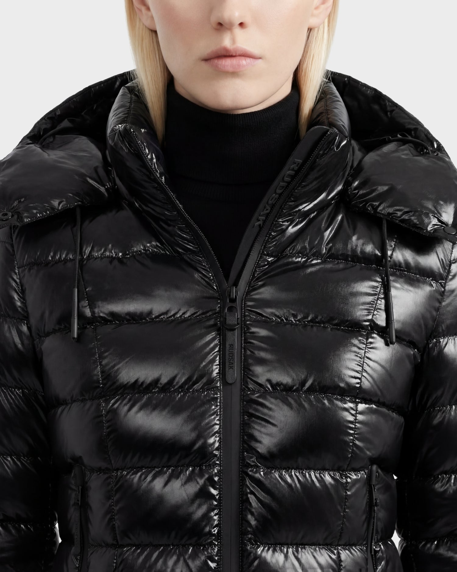 A person wearing the Rudsak MELISSA C - 8125695 Black women's puffer jacket with a hood and a black turtleneck sweater, shown from the chest up against a plain background.