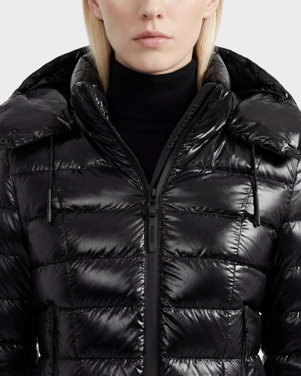 A person wearing the Rudsak MELISSA C - 8125695 Black women's puffer jacket with a hood and a black turtleneck sweater, shown from the chest up against a plain background.