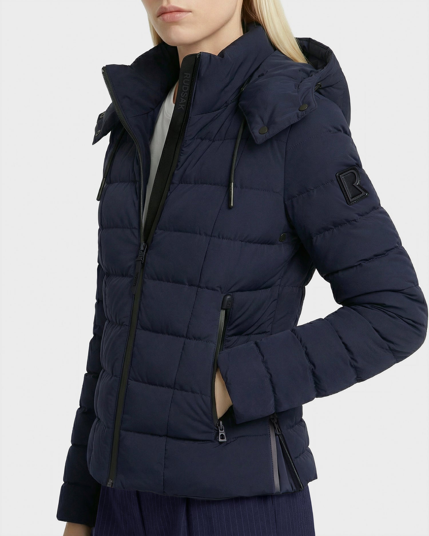 A woman in the Rudsak MELISSA - 8125054 Navy puffer jacket with RDS certified down, black zippers, hood, and upper sleeve patch stands against a plain background.