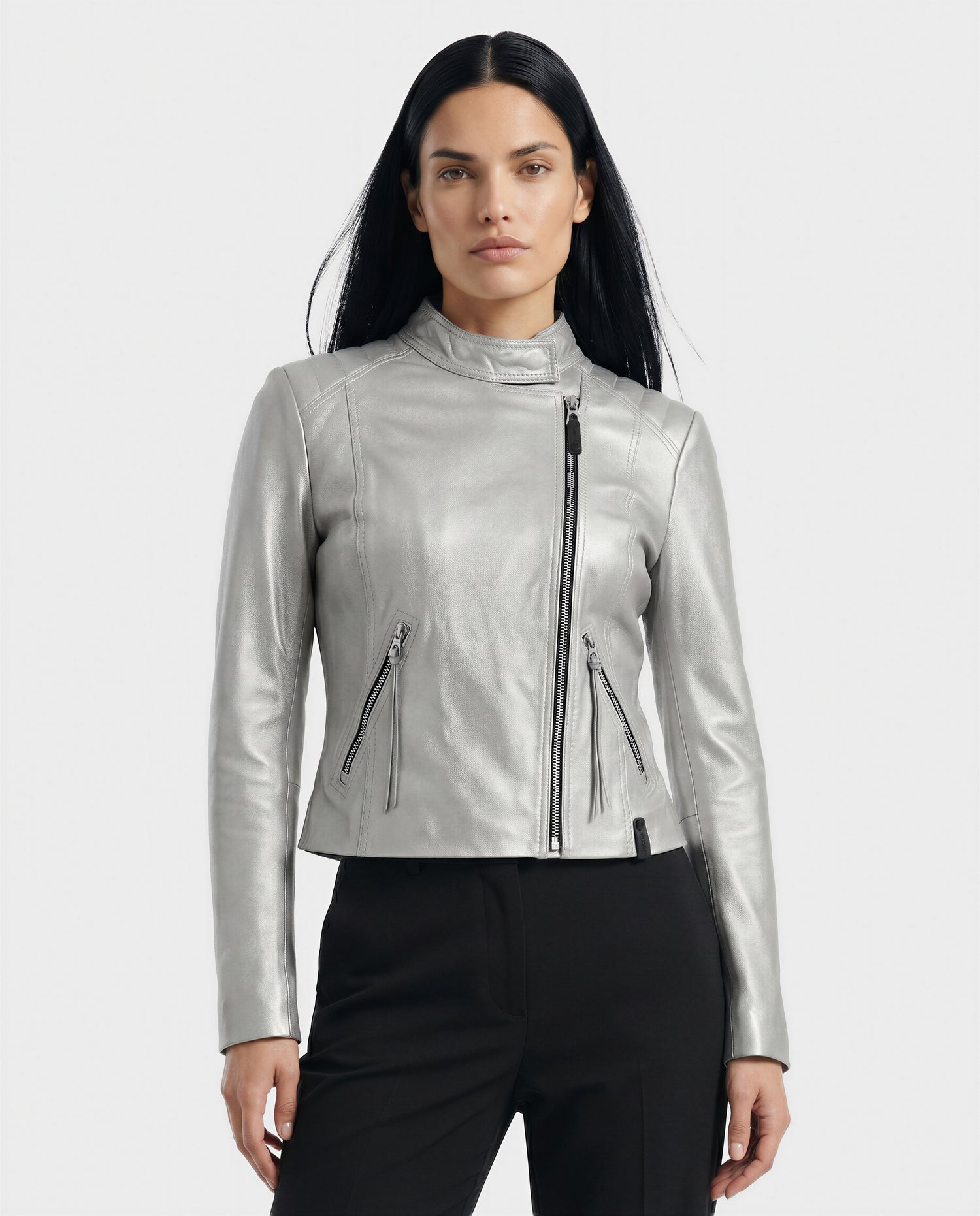 A woman with long black hair wears the Rudsak MERINA - L120013 Metallic Aluminium moto jacket with zippered pockets and an asymmetrical zipper, paired with black pants, standing against a plain light background.