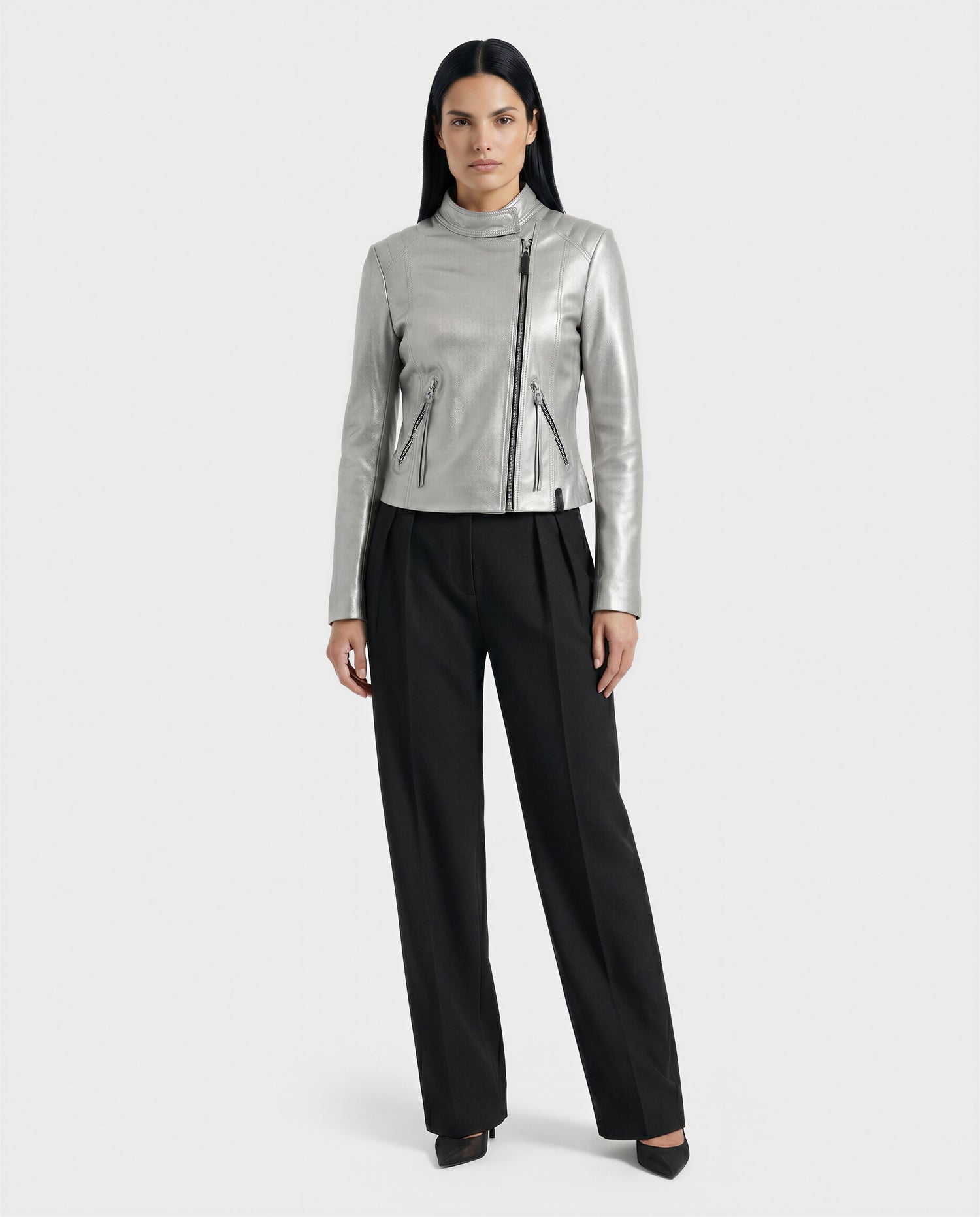A woman faces forward against a white background, wearing the Rudsak MERINA - L120013 Metallic Aluminium moto jacket with zipper details, paired with black pleated trousers and black heels.