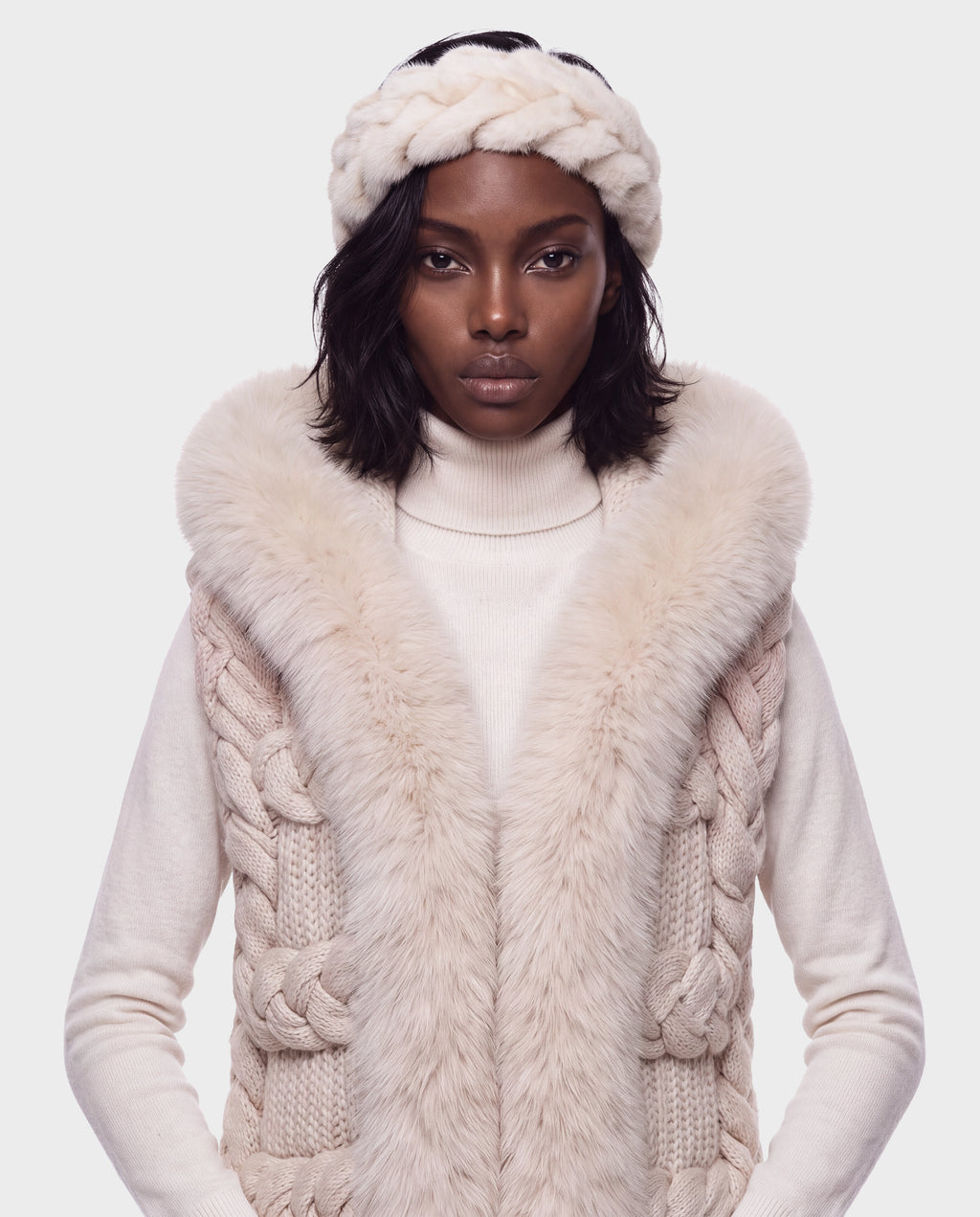 A person wearing a MILA - 8426518 Vanilla cable-knit vest with a fur-trimmed collar over a white turtleneck sweater, paired with a matching braided knit headband.