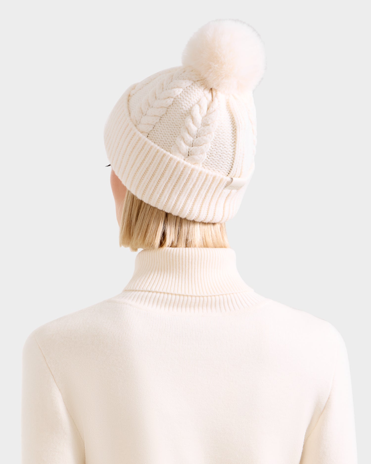 A person with short blonde hair, seen from behind, wears the Rudsak MAE - 8425550 Vanilla women’s cable knit tuque with faux fur pom pom and a matching cream turtleneck sweater against a light background.