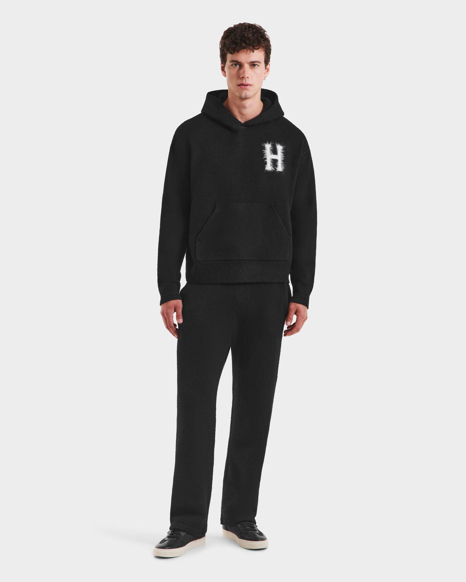 A person faces forward in a collegiate style, wearing Rudsak’s MARCUS HV - H525503 Black hoodie and matching pants, with a white “H” on the chest. They pair the look with black sneakers and have short curly hair, all set against a plain white background.