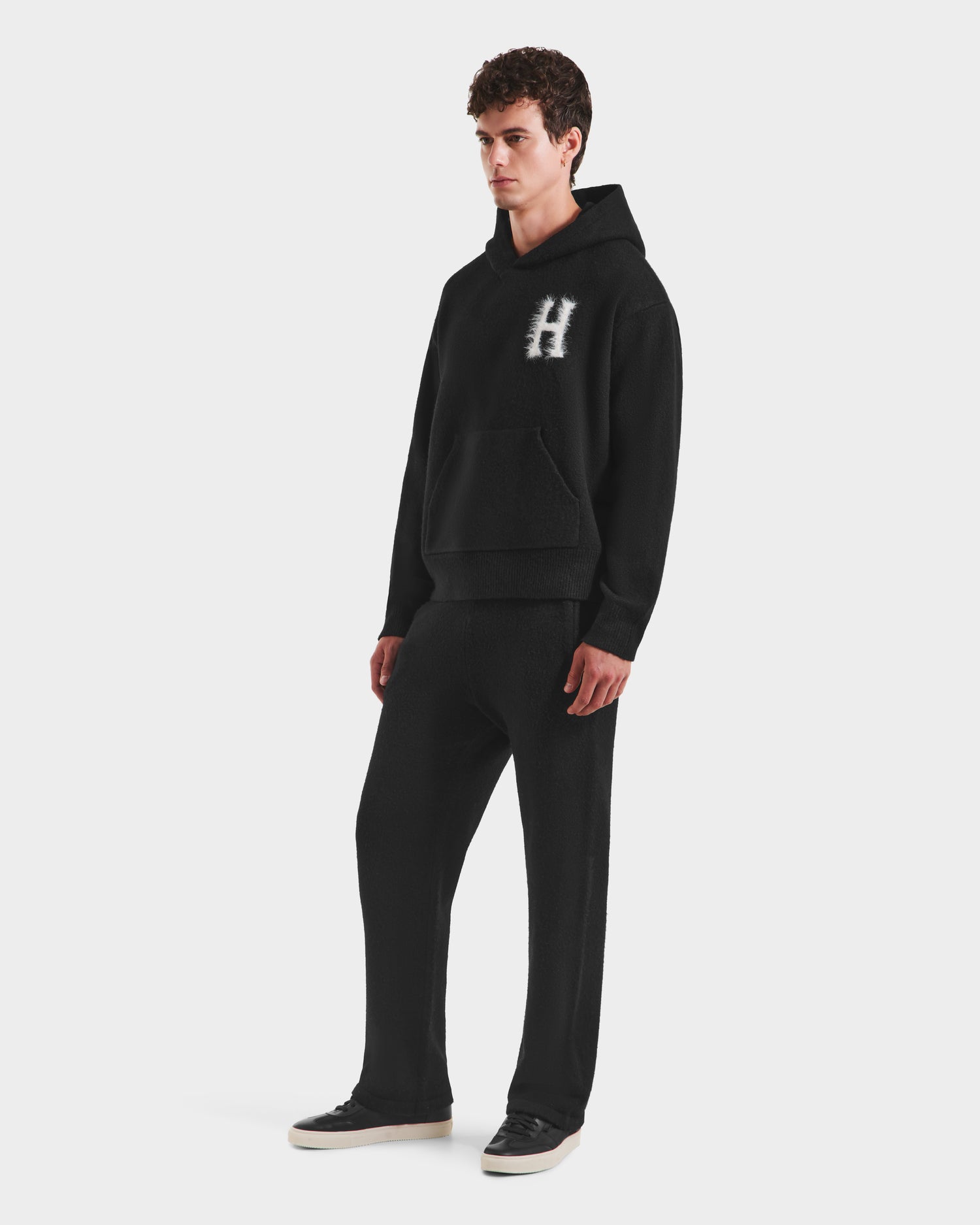 A young man poses against a white background in the Rudsak MARCUS HV - H525503 Black hoodie, featuring a bold white "H" on the chest, paired with matching black pants and sneakers for a classic collegiate look.