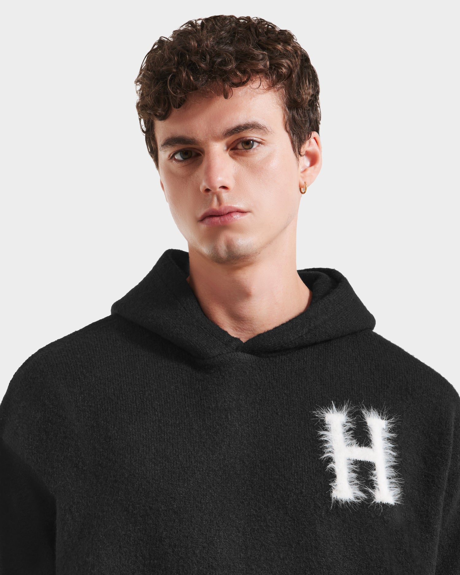 A young man with short curly brown hair and a small hoop earring wears the Rudsak MARCUS HV - H525503 Black collegiate hoodie, featuring a white fuzzy letter H on the left chest. He has a neutral expression.