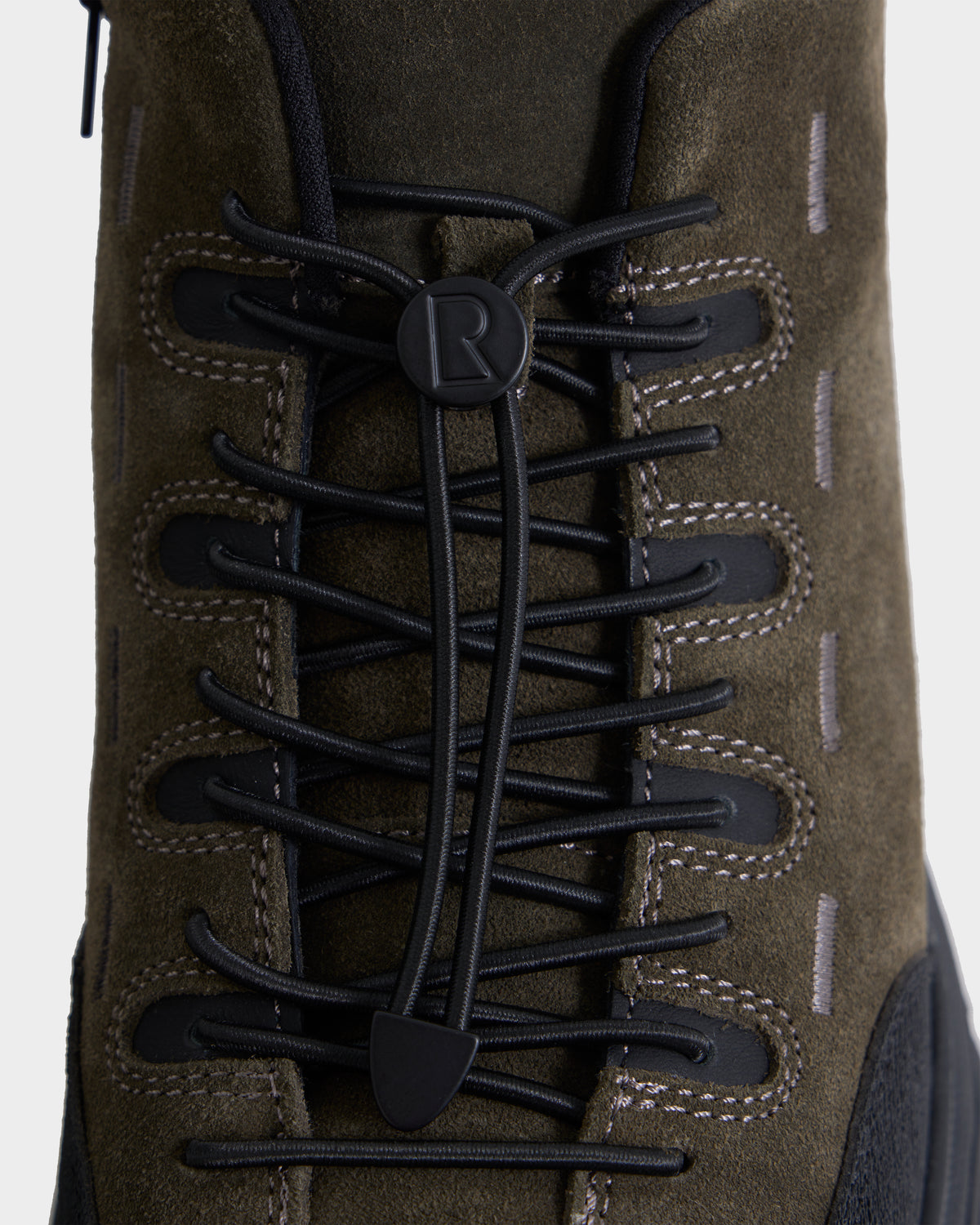 Close-up of the MAX S - 6226505 Olive boot by Rudsak in olive green suede, featuring black laces threaded through plastic lace hooks and accented with visible white stitching. Trail-inspired design elements include rugged construction and a signature black toggle embossed with the letter “R,” combining classic boot style with a modern edge.