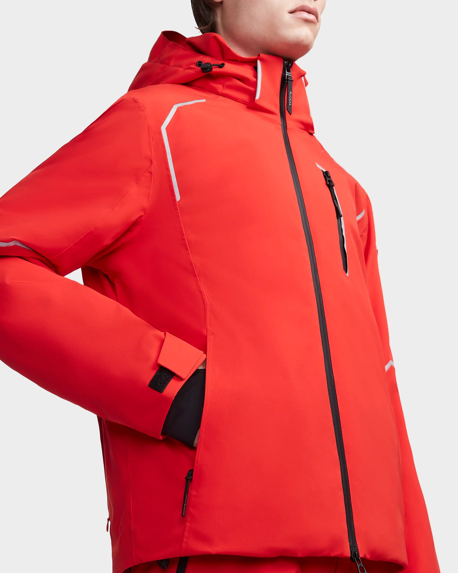 A person wears the Rudsak EDMONTON - 6123520 Orange men’s ski jacket with black zipper details and white accents. This outdoor winter jacket features a high collar, zippered pockets, and may include a RECCO Reflector for added safety.