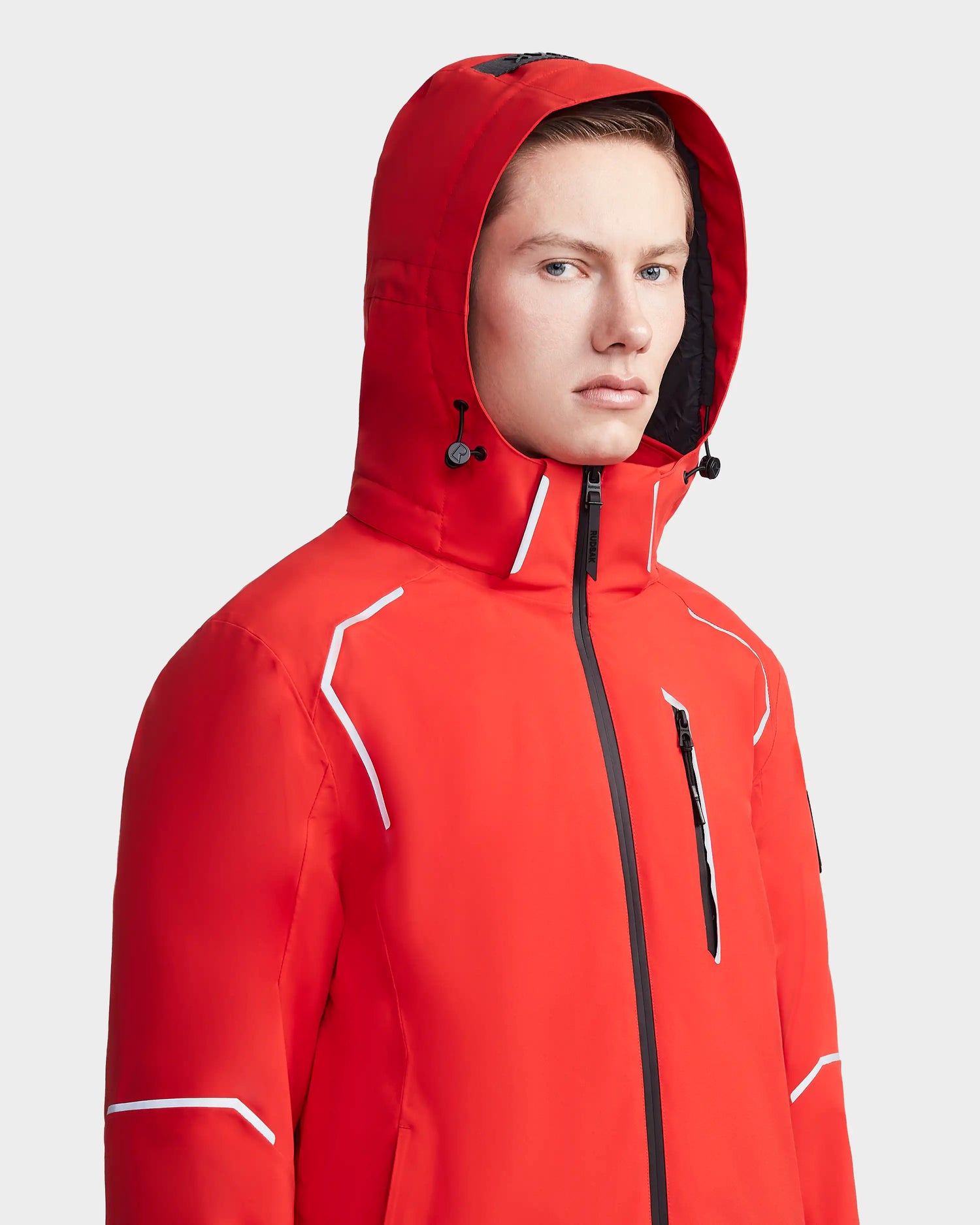 A person with light skin and short hair wears the Rudsak EDMONTON - 6123520 Orange men's ski jacket, featuring white accents, a zippered chest pocket, and RECCO Reflector, looking forward neutrally against a plain light background.