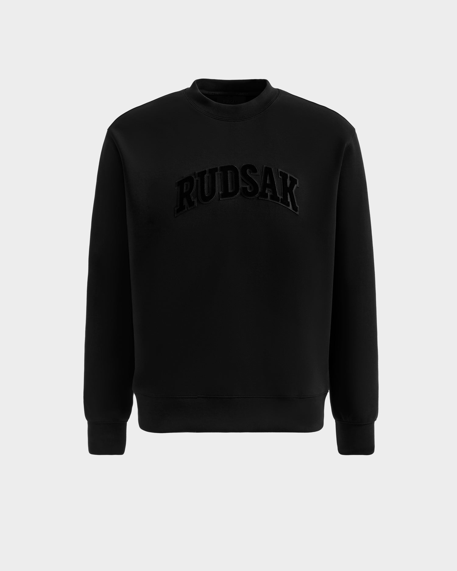 The Rudsak MILOS - 6525646 Black is a men's premium crewneck sweatshirt with long sleeves, made from a soft cotton blend and featuring bold raised RUDSAK lettering across the chest, shown against a plain light grey background.