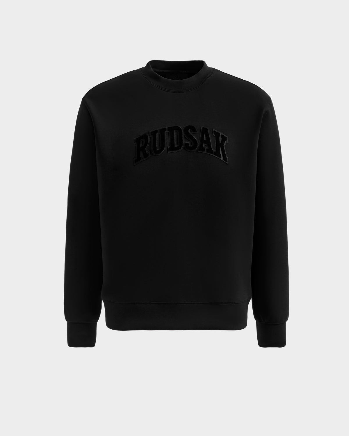 The Rudsak MILOS - 6525646 Black is a men's premium crewneck sweatshirt with long sleeves, made from a soft cotton blend and featuring bold raised RUDSAK lettering across the chest, shown against a plain light grey background.