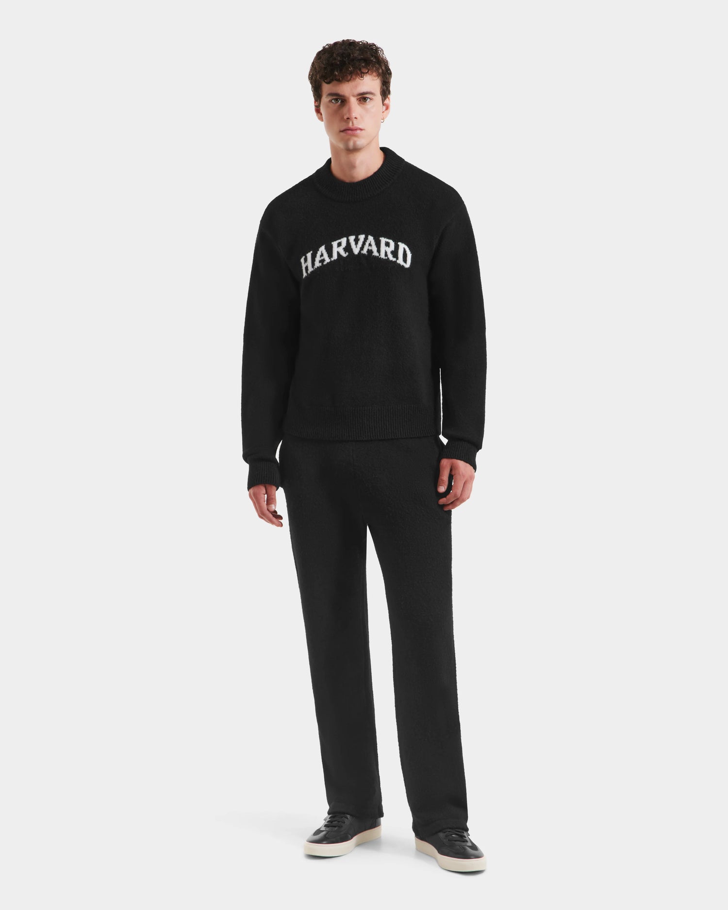 A young man stands against a plain background wearing the limited-edition Rudsak NEVE HV - H525504 Black sweater with white lettering, paired with matching black pants and black sneakers.