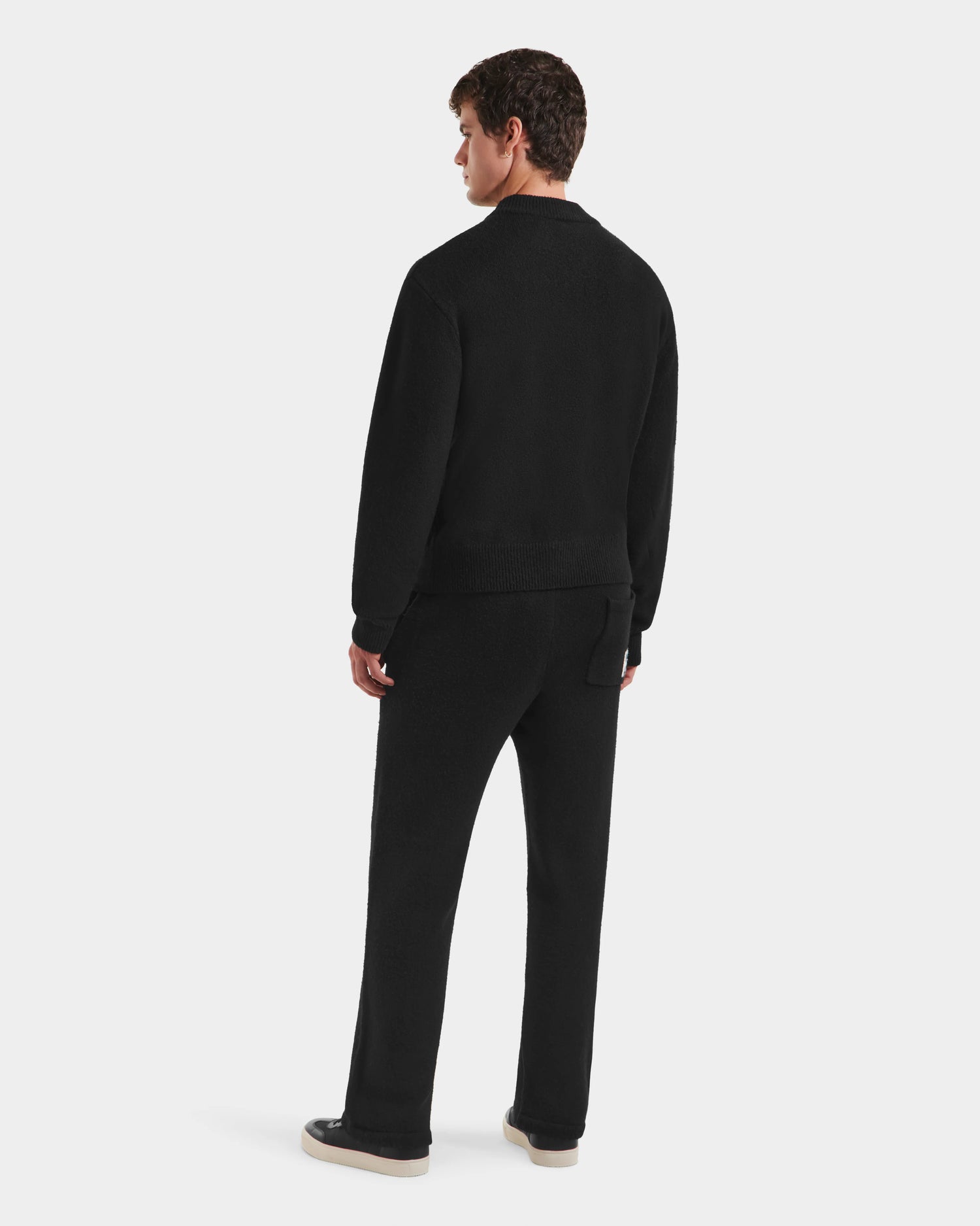 A man faces away in the Rudsak NEVE HV - H525504 Black knit, styled with black pants and beige-soled black shoes, all set against a plain white background.