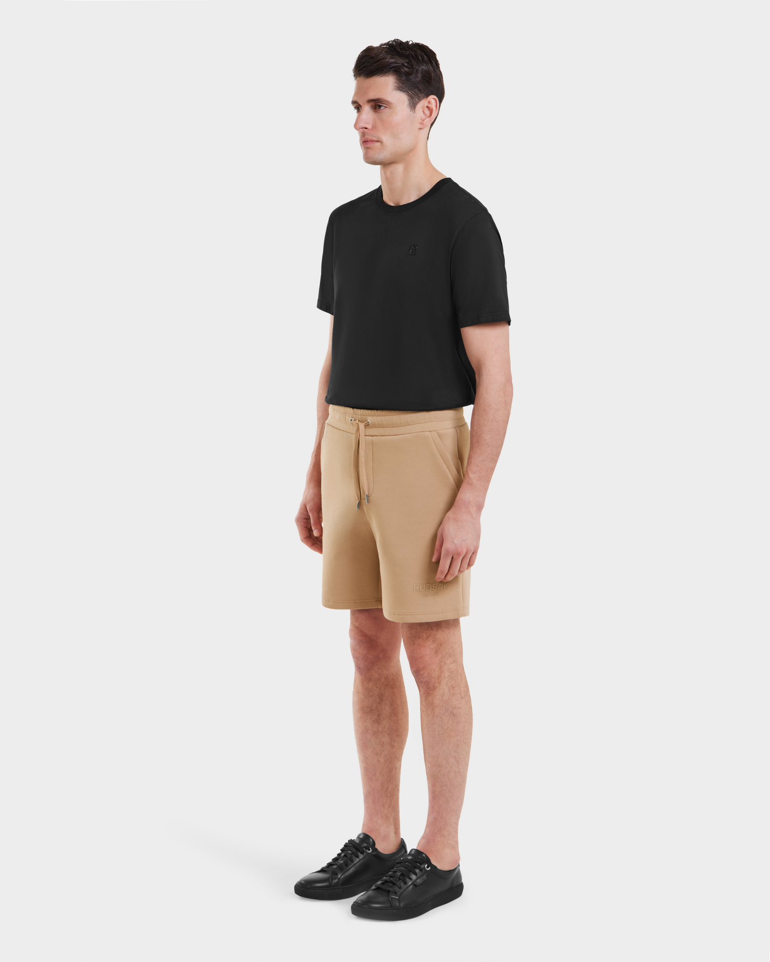 A man stands against a white background, wearing Rudsak PASCAL - 6524022 Almond shorts with a stretch waistband, a black t-shirt, and black sneakers. He faces slightly left with arms relaxed at his sides.