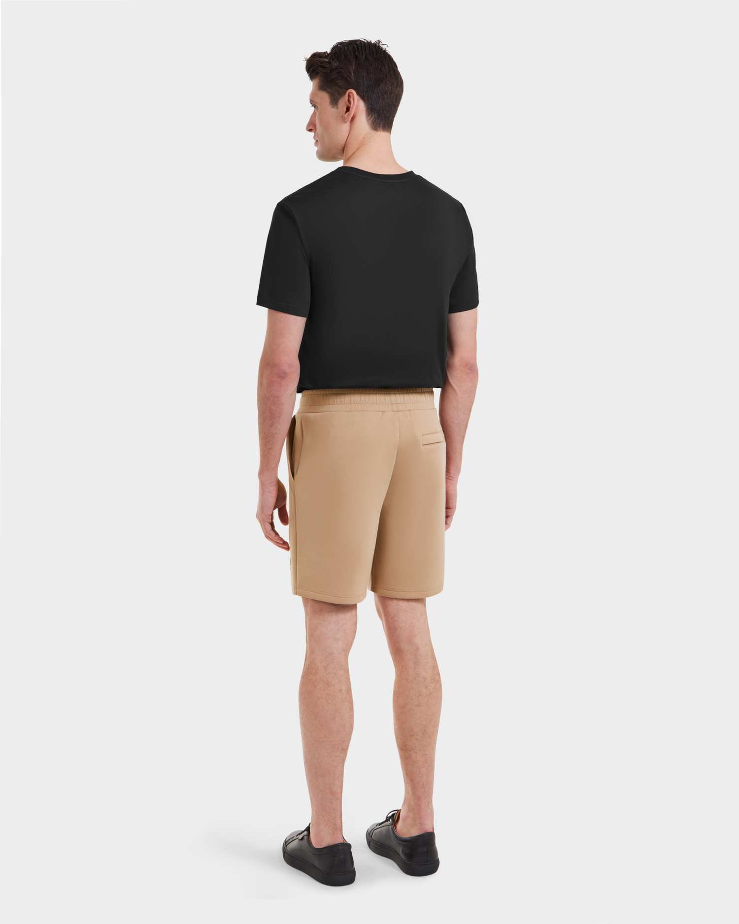 A man stands facing away, wearing the Rudsak PASCAL - 6524022 Almond shorts with a stretch waistband, a black short-sleeve t-shirt, and black shoes against a plain white background.
