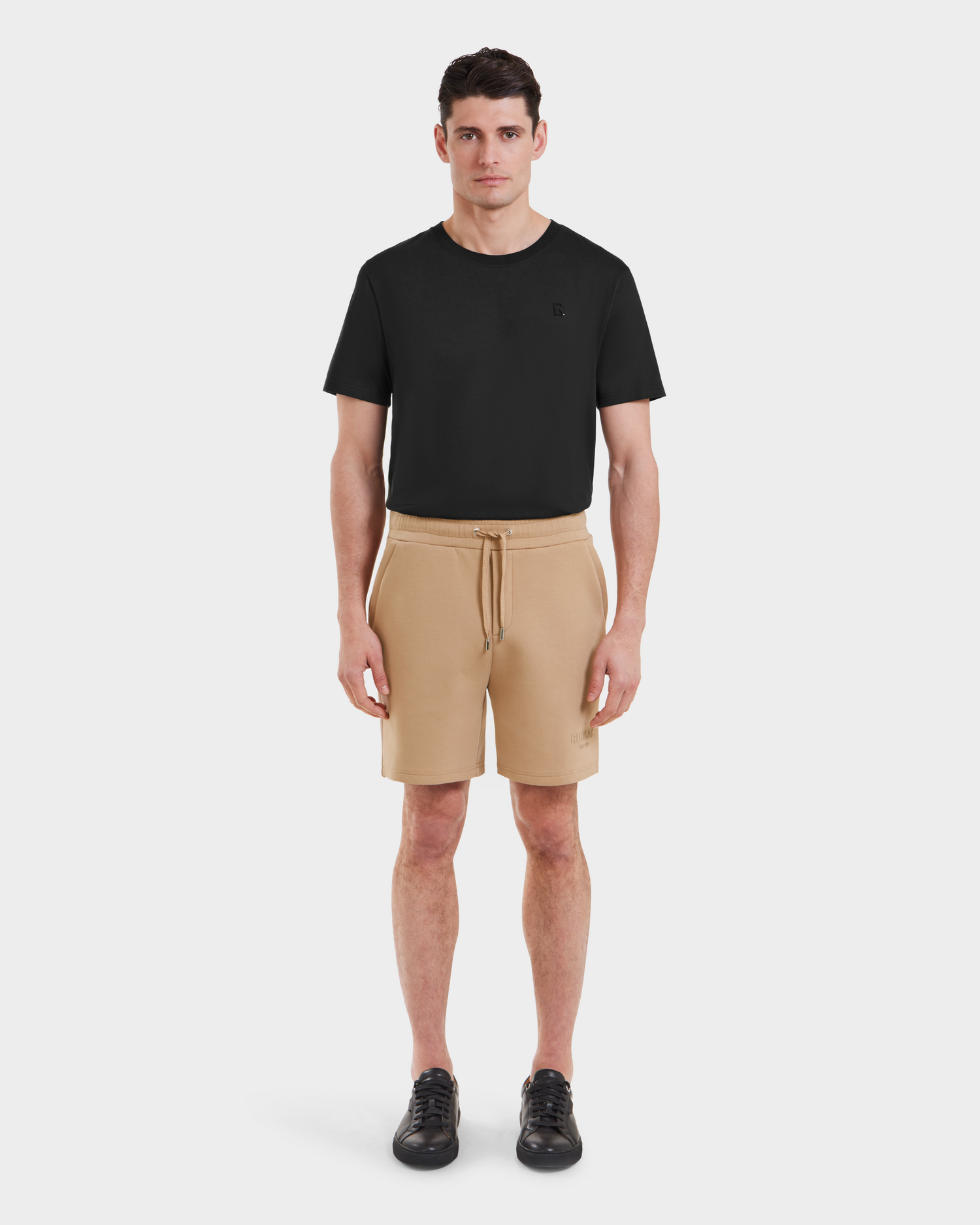 A man stands facing forward, wearing a black short-sleeve t-shirt, Rudsak PASCAL - 6524022 Almond stretch waistband shorts, and black sneakers against a plain white background.