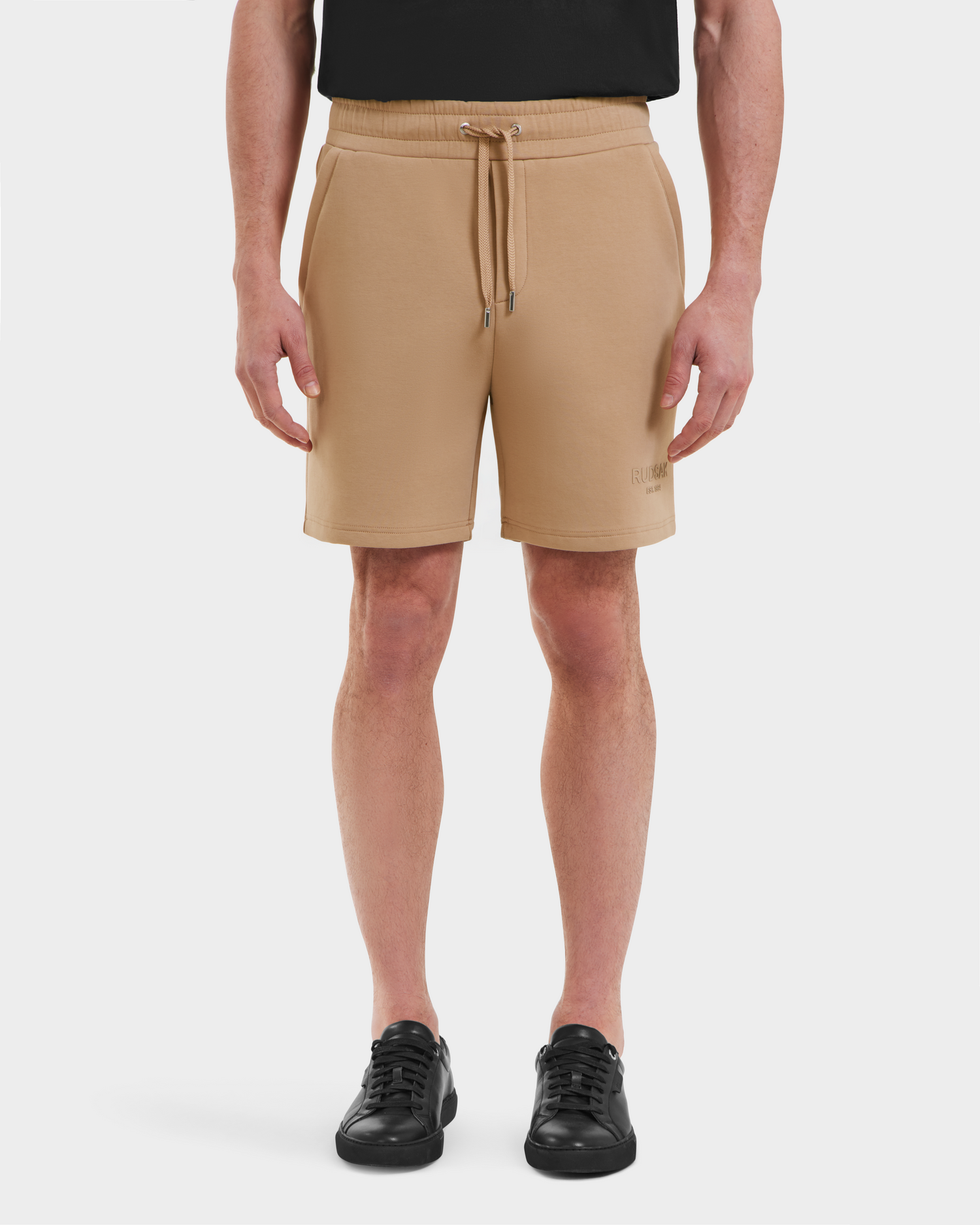 A person faces forward against a plain white background, wearing Rudsak's PASCAL - 6524022 Almond shorts with a stretch waistband, paired with black sneakers and a partially visible black shirt.