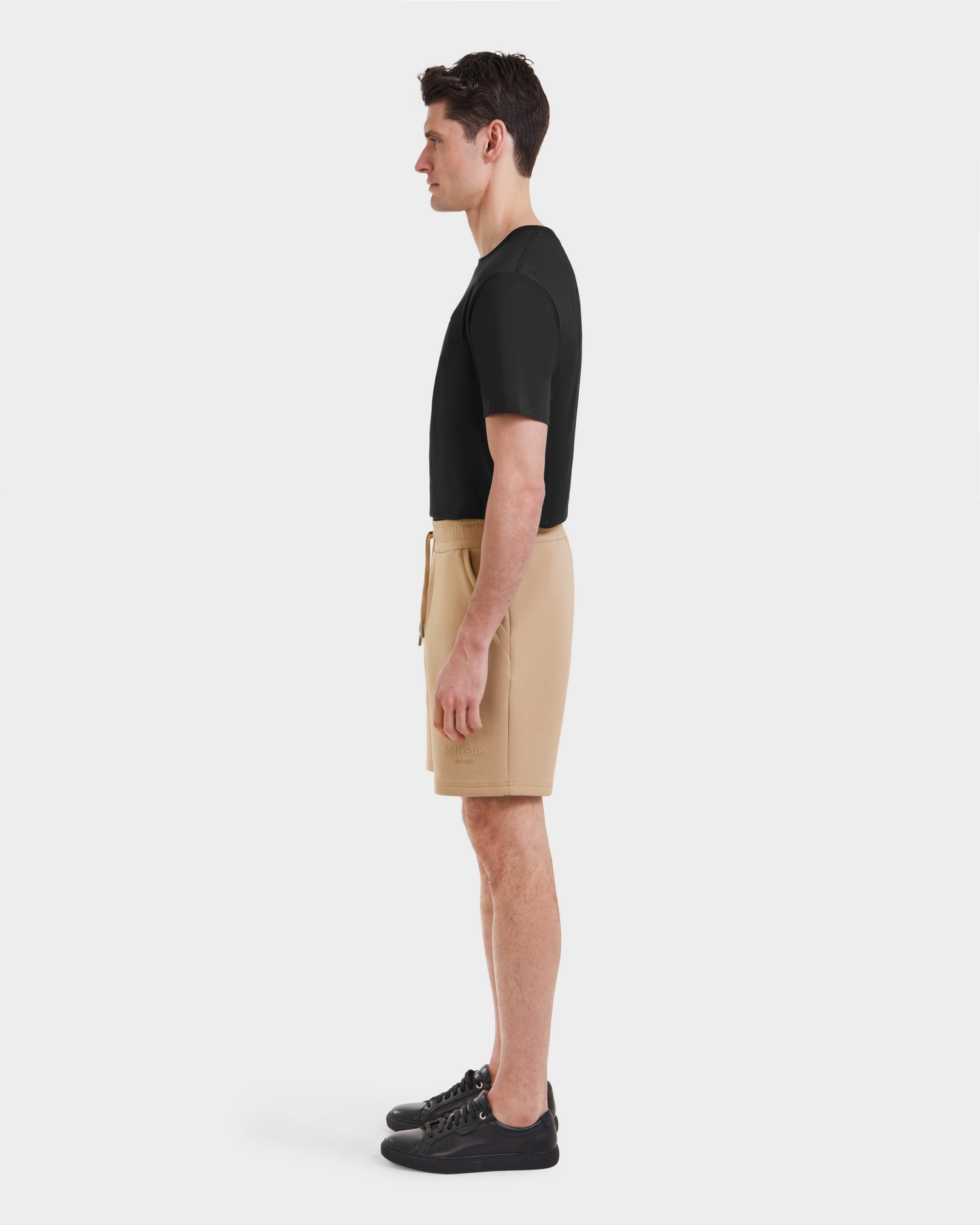 A man stands in profile facing left, wearing a black t-shirt, Rudsak PASCAL - 6524022 Almond shorts, and black sneakers, against a plain white background.