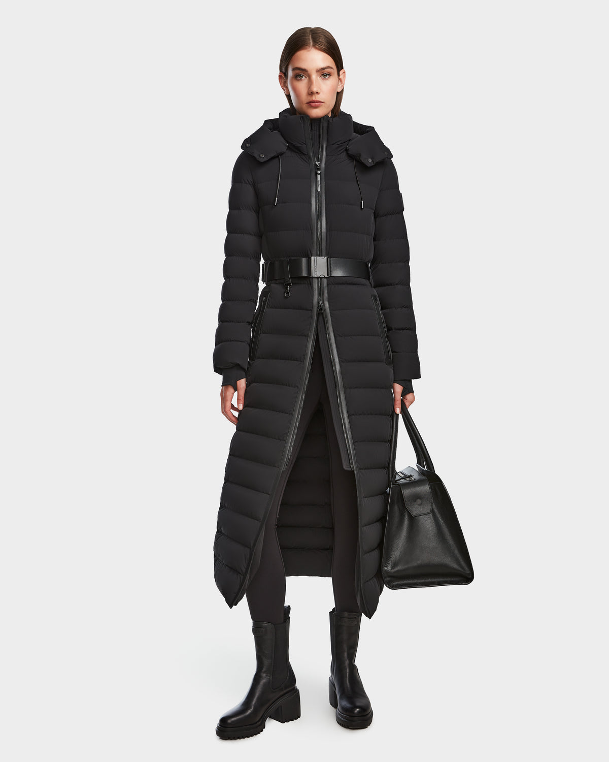 A woman faces forward in the Rudsak PELIN L - 8124815 Black womens long down puffer winter coat with a belt, paired with black leggings, chunky boots, and a large black handbag against a plain white background.