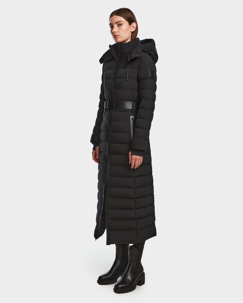 A woman wears the Rudsak PELIN L - 8124815 Black womens long down puffer winter coat with hood and black zipper, paired with black boots. The belted coat is water- and wind-resistant, shown against a plain white background.