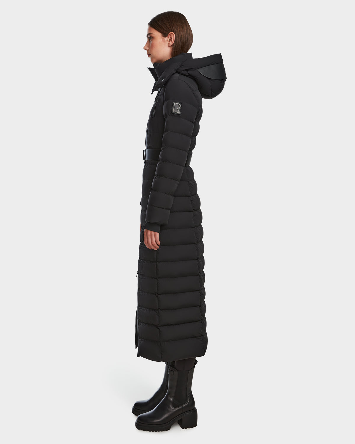 A woman stands in profile against a white background, wearing the Rudsak PELIN L - 8124815 Black womens long down puffer winter coat with hood, which is water- and wind-resistant, paired with black heeled boots.