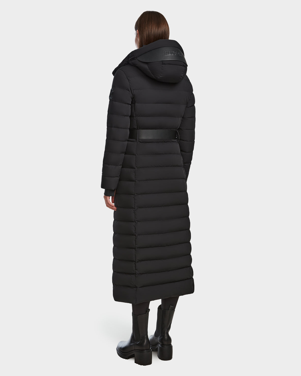 A person stands facing away from the camera in the Rudsak PELIN L - 8124815 Black womens long down puffer winter coat with hood and belt, paired with black pants and boots. The coat is water and wind-resistant for style and protection.
