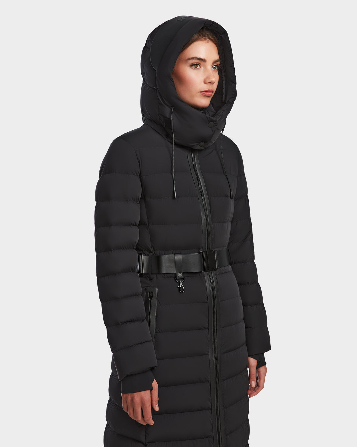 A woman wearing the Rudsak PELIN L - 8124815 Black womens long down puffer coat with a hood and wide belt stands against a plain white background. This winter coat is water- and wind-resistant for cold-weather protection.