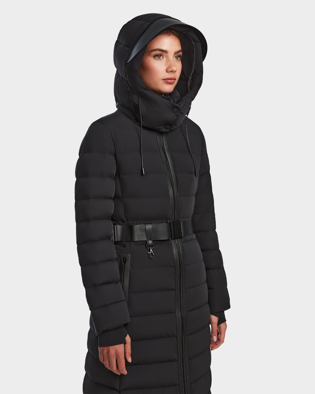 A woman stands against a white background wearing the Rudsak PELIN L - 8124815 Black womens long down puffer with hood and belted waist. This stylish coat offers water and wind resistance for added protection.