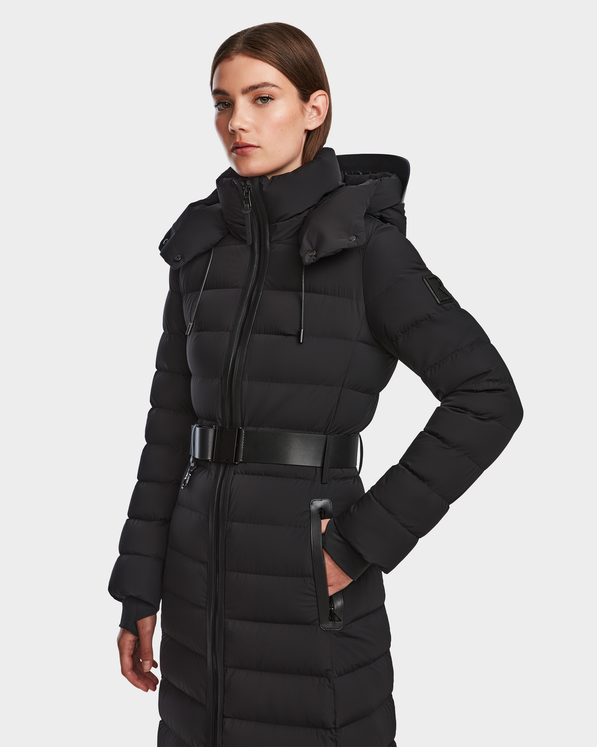 A woman models the Rudsak PELIN L - 8124815 Black womens long down puffer featuring a hood and belted waist, standing slightly sideways against a plain white background.