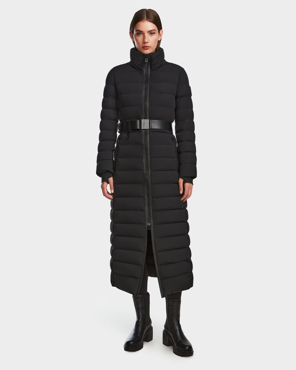 A woman faces forward in the Rudsak PELIN L - 8124815 Black womens long down puffer winter coat with a belted waist, black pants, and boots. The stylish water- and wind-resistant coat is perfect for cold weather against a white background.