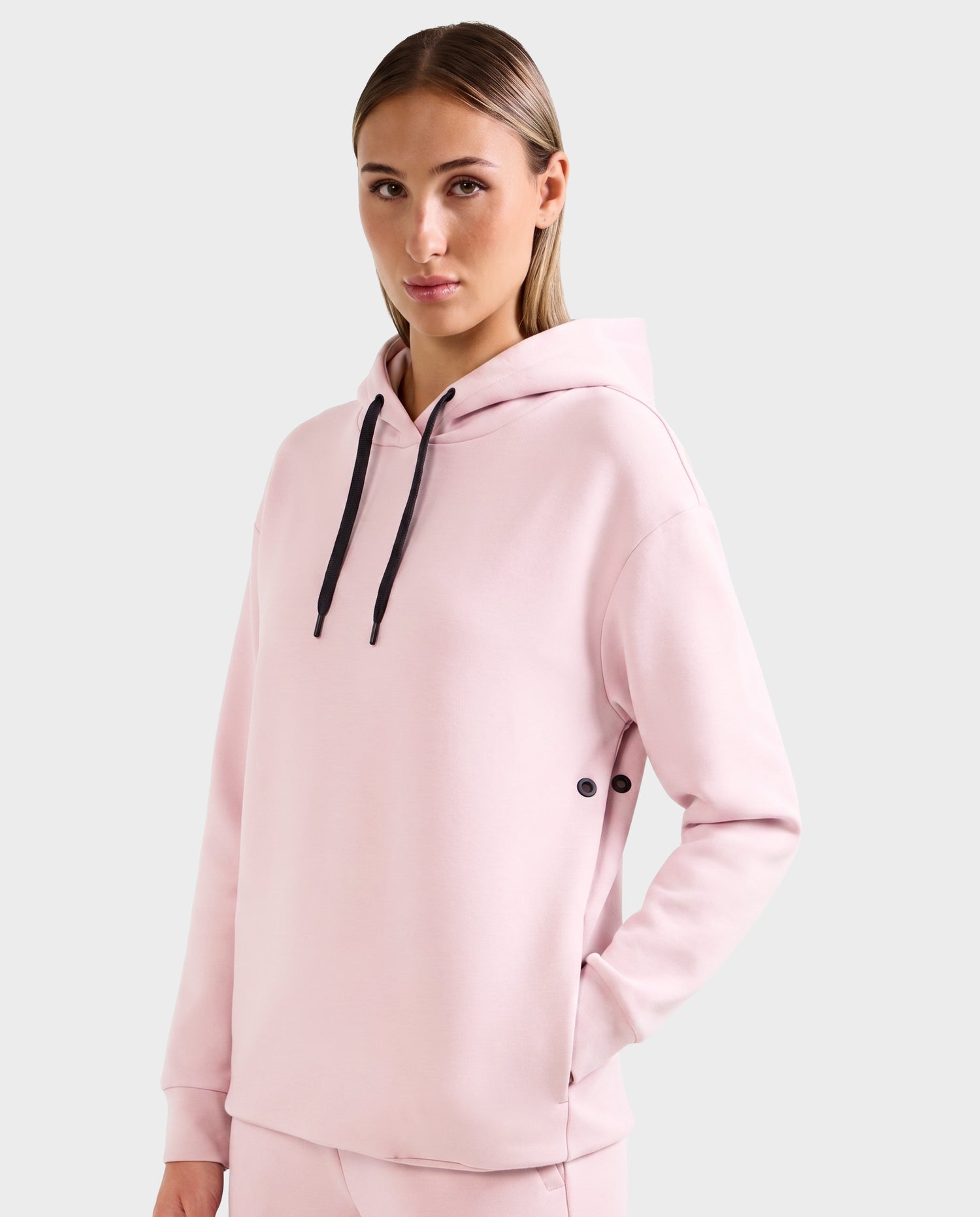 A person wears the Rudsak LEROY - U525509 Light Pink W unisex hoodie with black drawstrings and matching pants, standing against a light gray background and looking at the camera with a neutral expression.