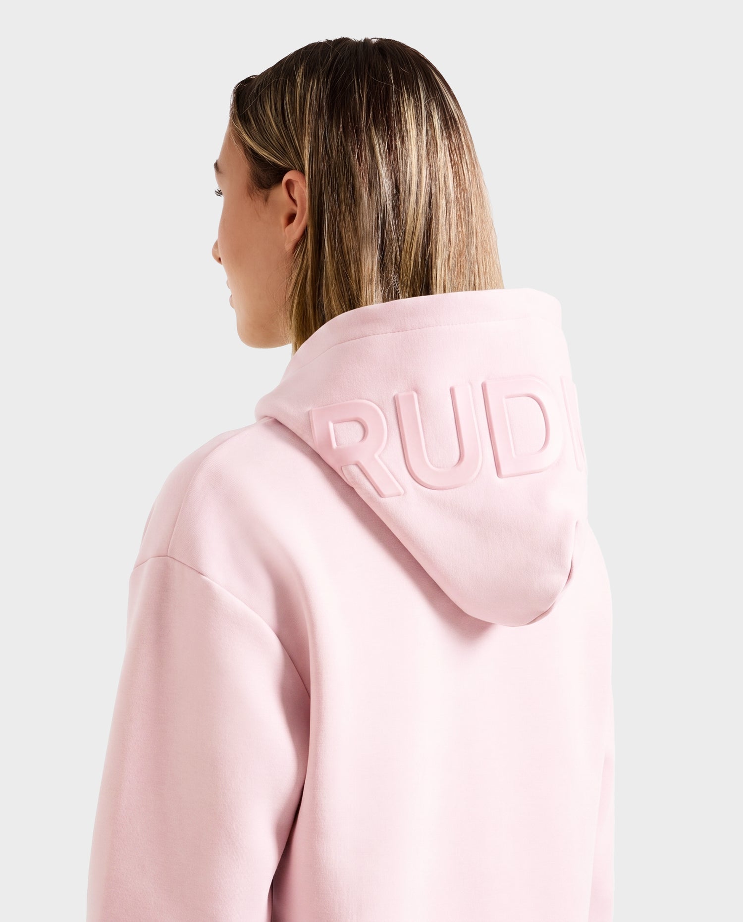 A woman with straight, light brown hair wears the Rudsak LEROY - U525509 Light Pink W premium hoodie, featuring embossed "RUDI" text on the hood. She is turned away from the camera against a plain light gray background.