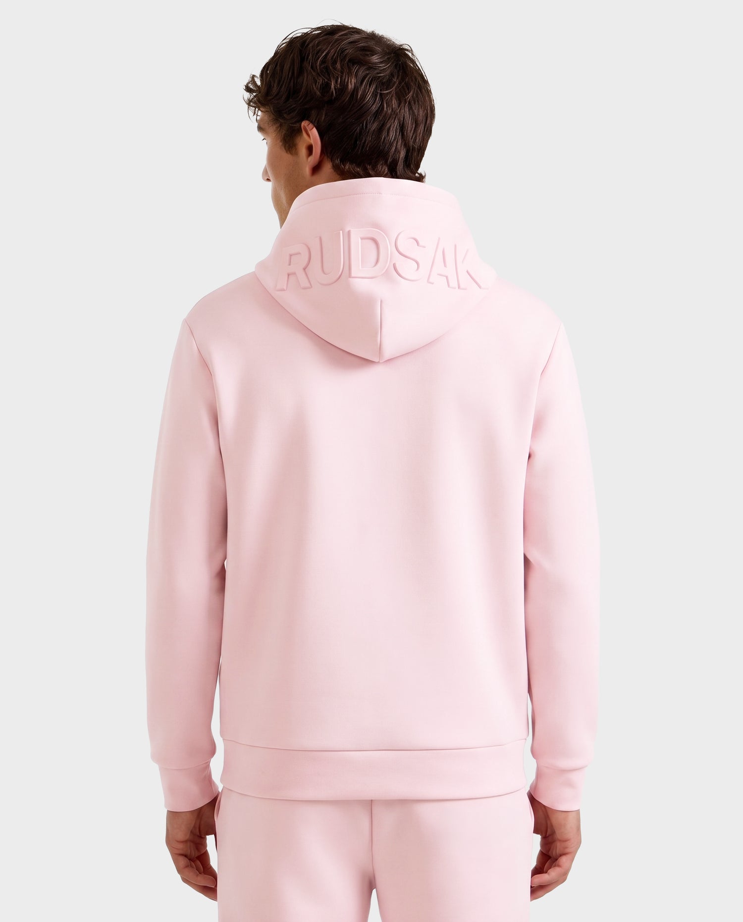 A person is seen from behind wearing the Rudsak LEROY - U525509 hoodie in light pink (size M) with the hood up and "RUDSAK" embossed on it, paired with matching pink pants.