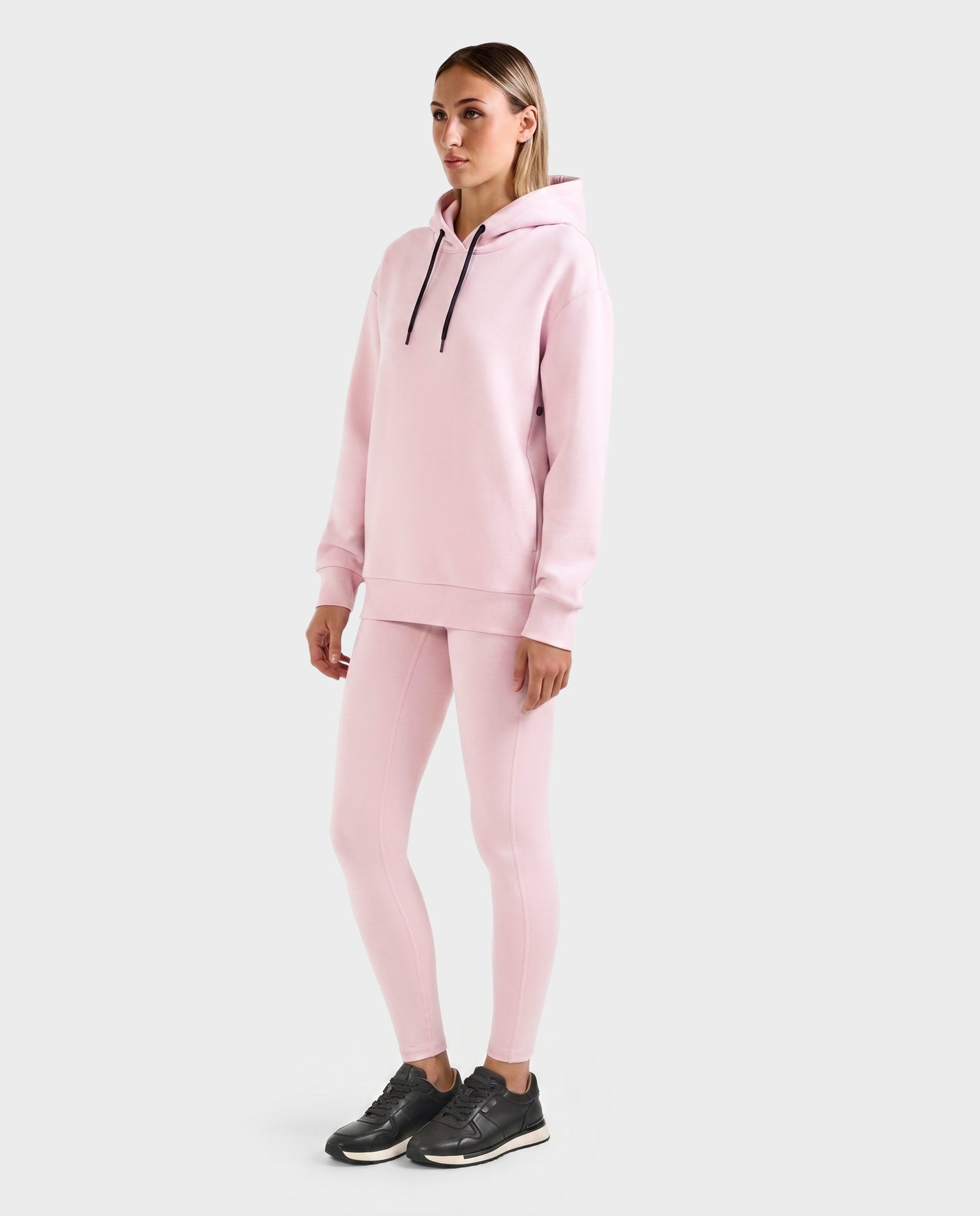 A person stands against a plain background wearing the Rudsak LEROY - U525509 Light Pink W gender-inclusive hoodie and matching leggings, paired with black athletic shoes.