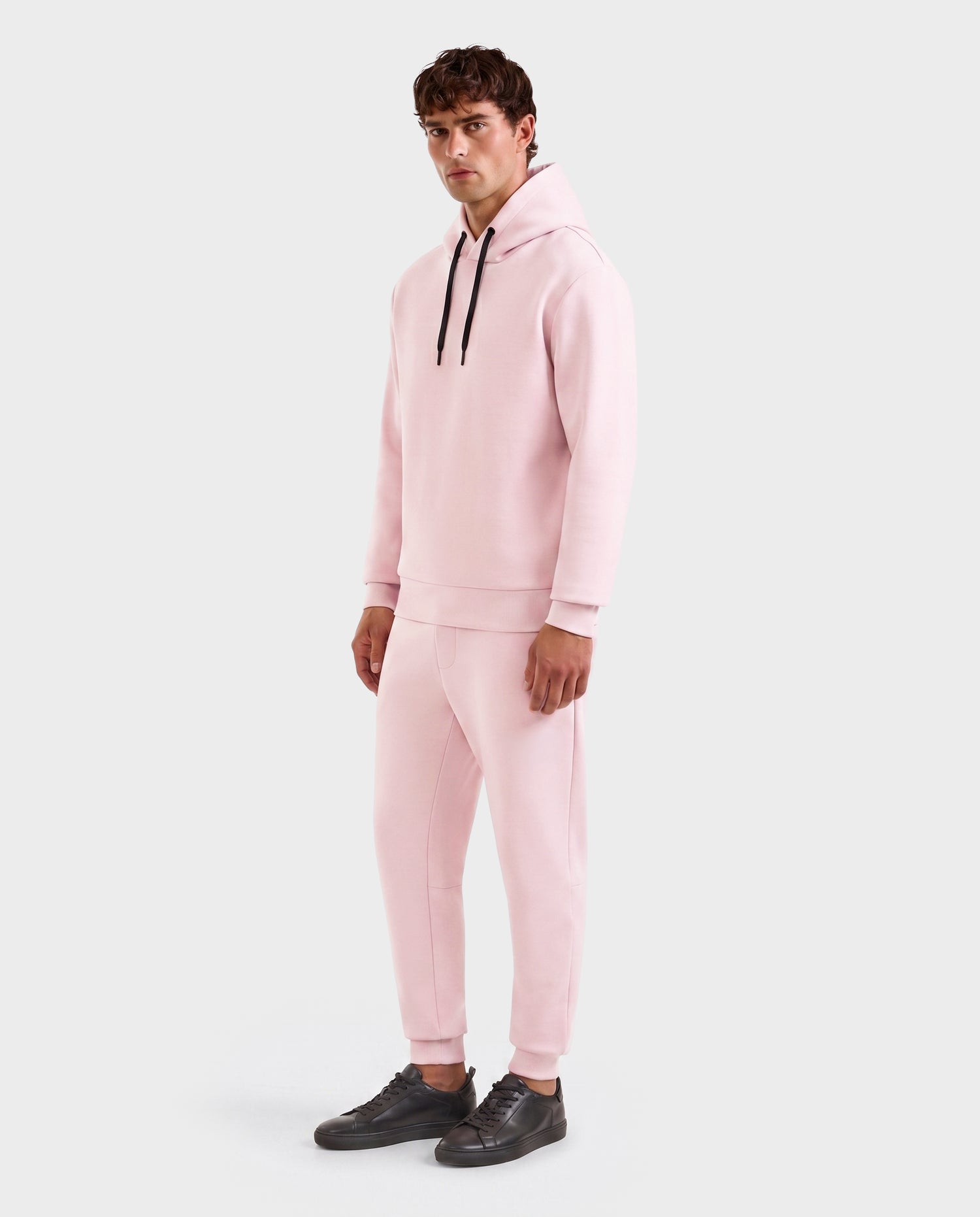 A person wears the Rudsak LEROY - U525509 Light Pink M unisex hoodie with black drawstrings and matching jogger pants, paired with black sneakers, standing against a plain light gray background.