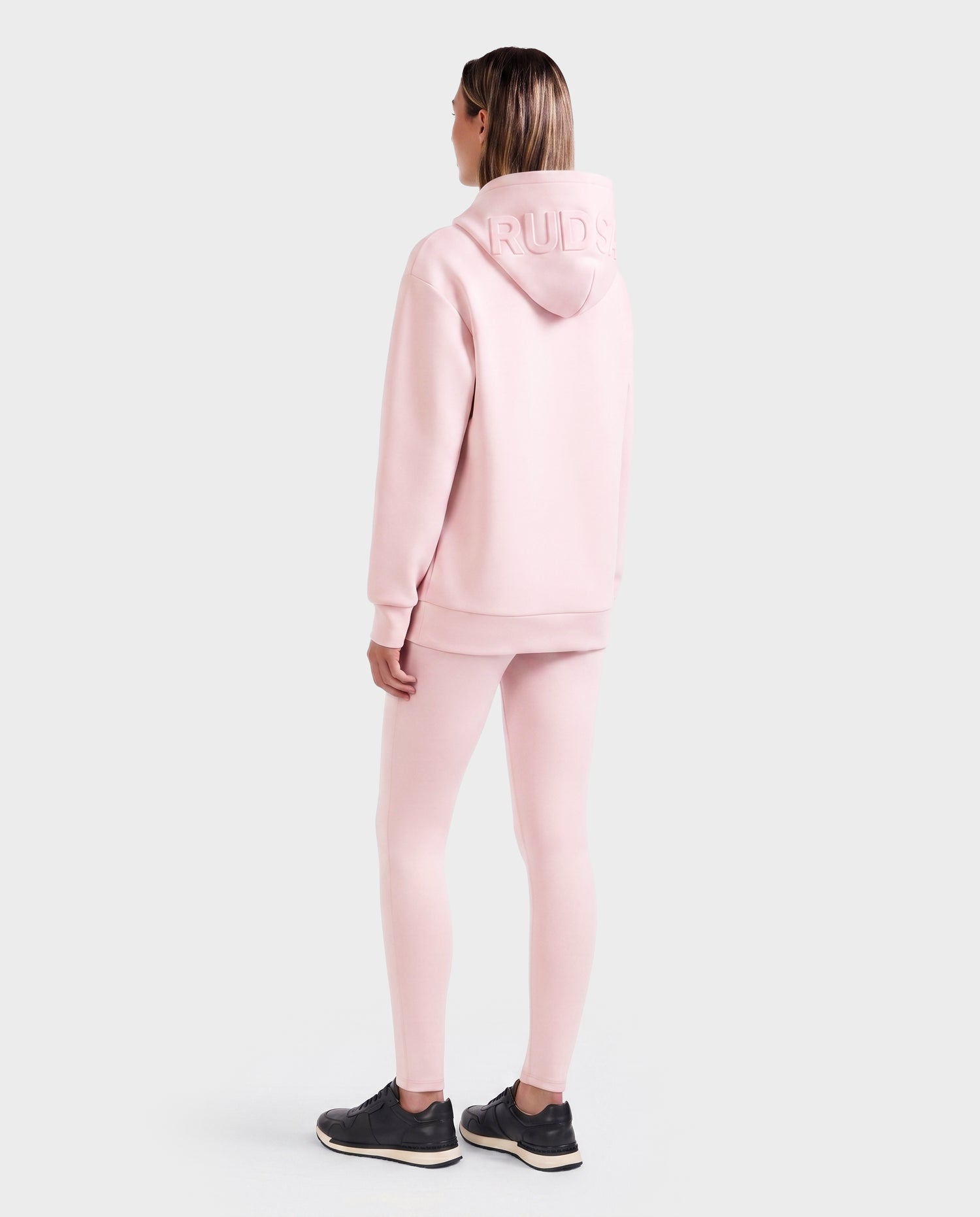A person faces away, wearing the Rudsak LEROY - U525509 Light Pink W gender-inclusive hoodie with “RUDS” on the hood and matching pink leggings. They have shoulder-length hair and black athletic shoes on a light gray background.