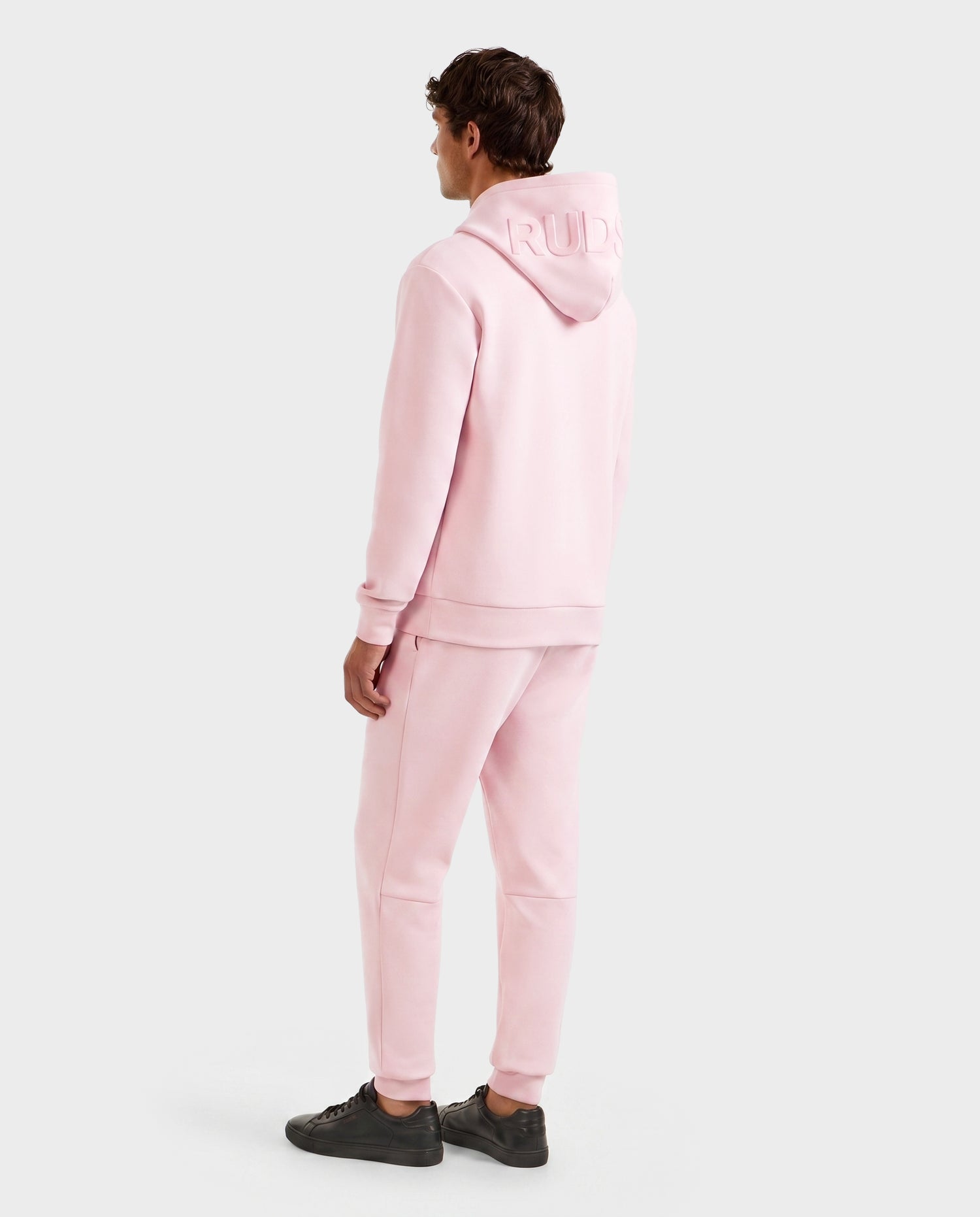 A person faces away, wearing the Rudsak LEROY - U525509 Light Pink M premium hoodie and matching sweatpants with black sneakers. Embossed text appears on the hood's back. The background is plain and light-colored.