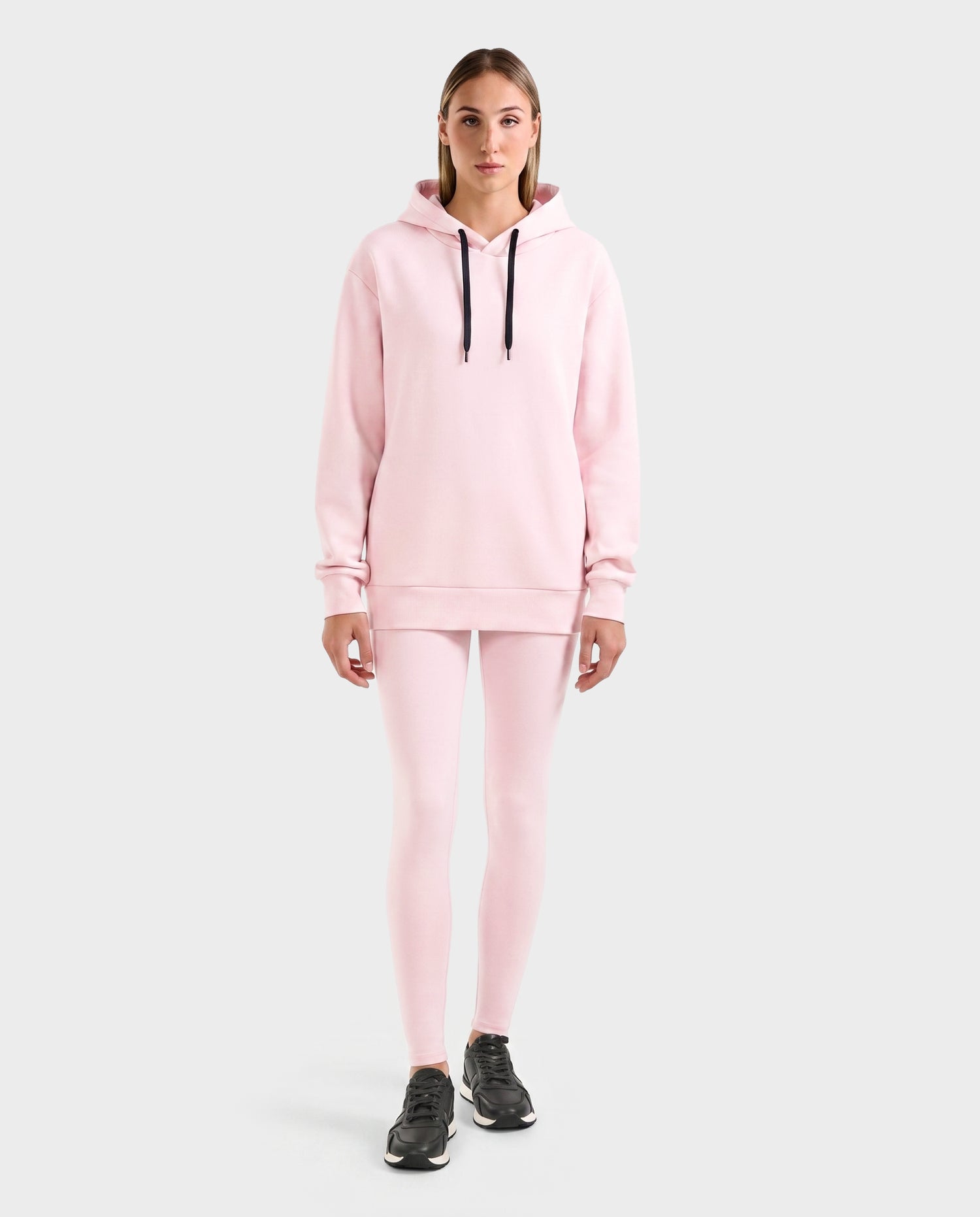 A person faces forward against a plain light gray background, wearing the Rudsak LEROY - U525509 Light Pink W hoodie with black drawstrings, matching pink leggings, and black athletic shoes.