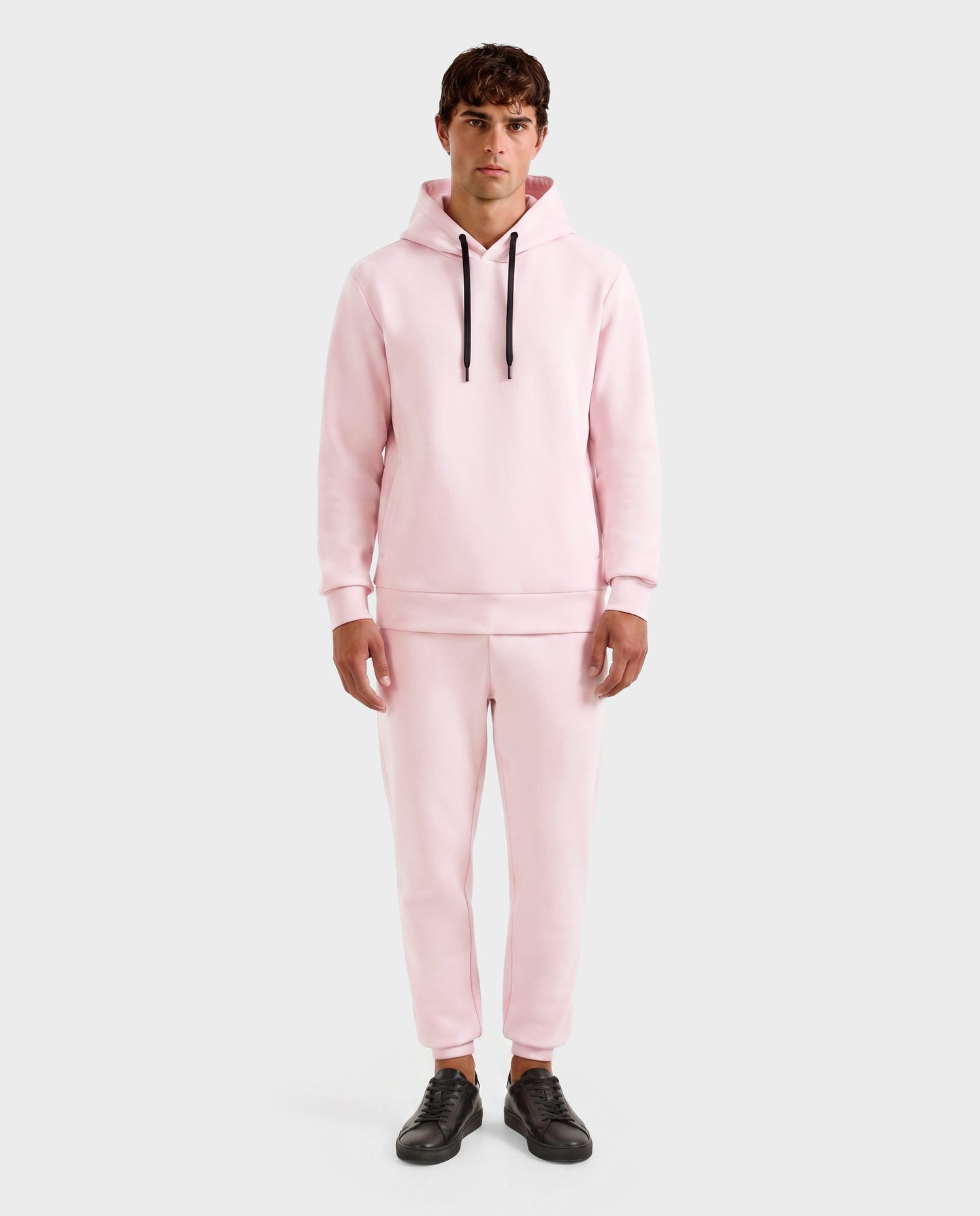 A man faces forward in a Rudsak LEROY - U525509 Light Pink M premium hoodie and jogger pants with black drawstrings, paired with black sneakers, set against a plain light gray background.