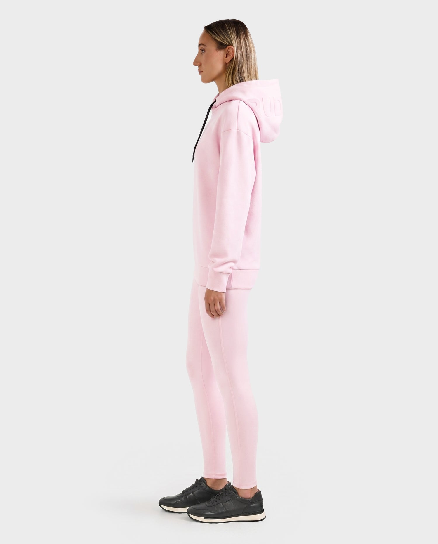 A woman in profile wears the Rudsak LEROY - U525509 Light Pink W hoodie and matching leggings with black sneakers, set against a plain light gray background.