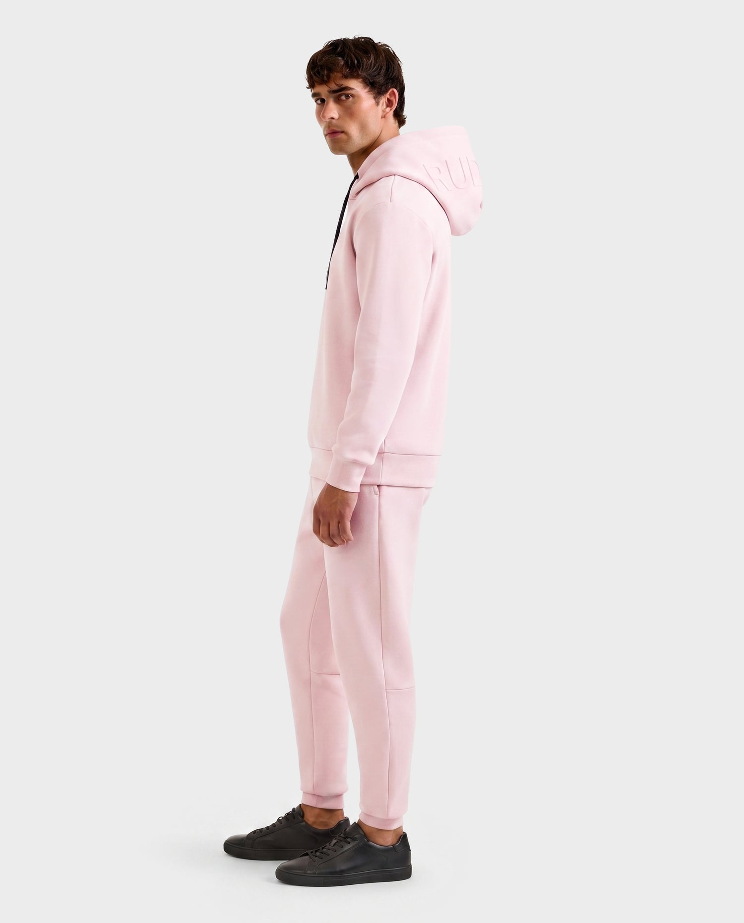 A man faces sideways to the camera, wearing the Rudsak LEROY - U525509 hoodie in light pink (size M) with matching joggers and black sneakers, set against a plain light gray background.
