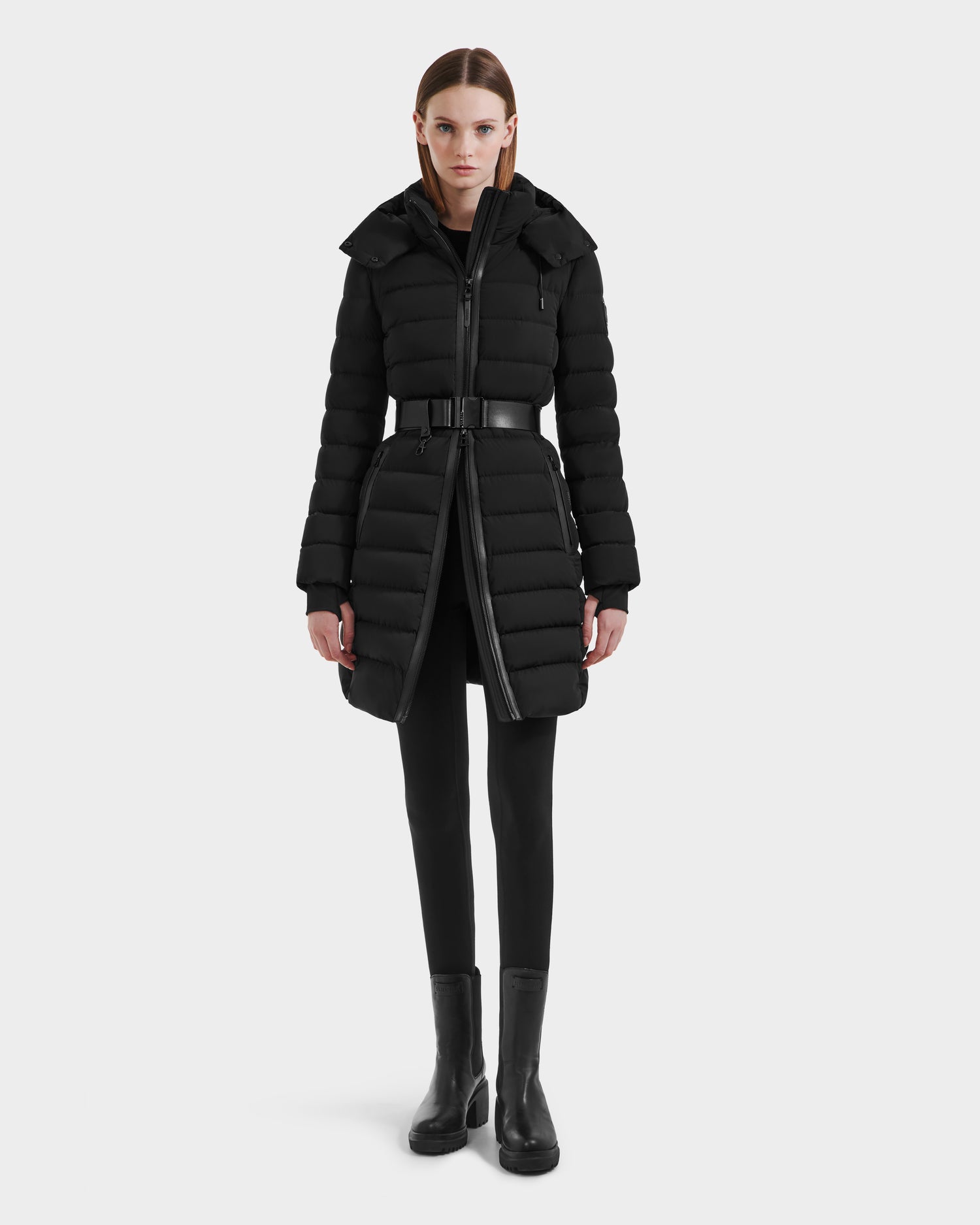 A woman faces forward in the Rudsak PELIN M HV - H125508 Black, a long black belted down jacket with vegan leather accents and high collar, styled with black leggings and ankle boots against a white background.