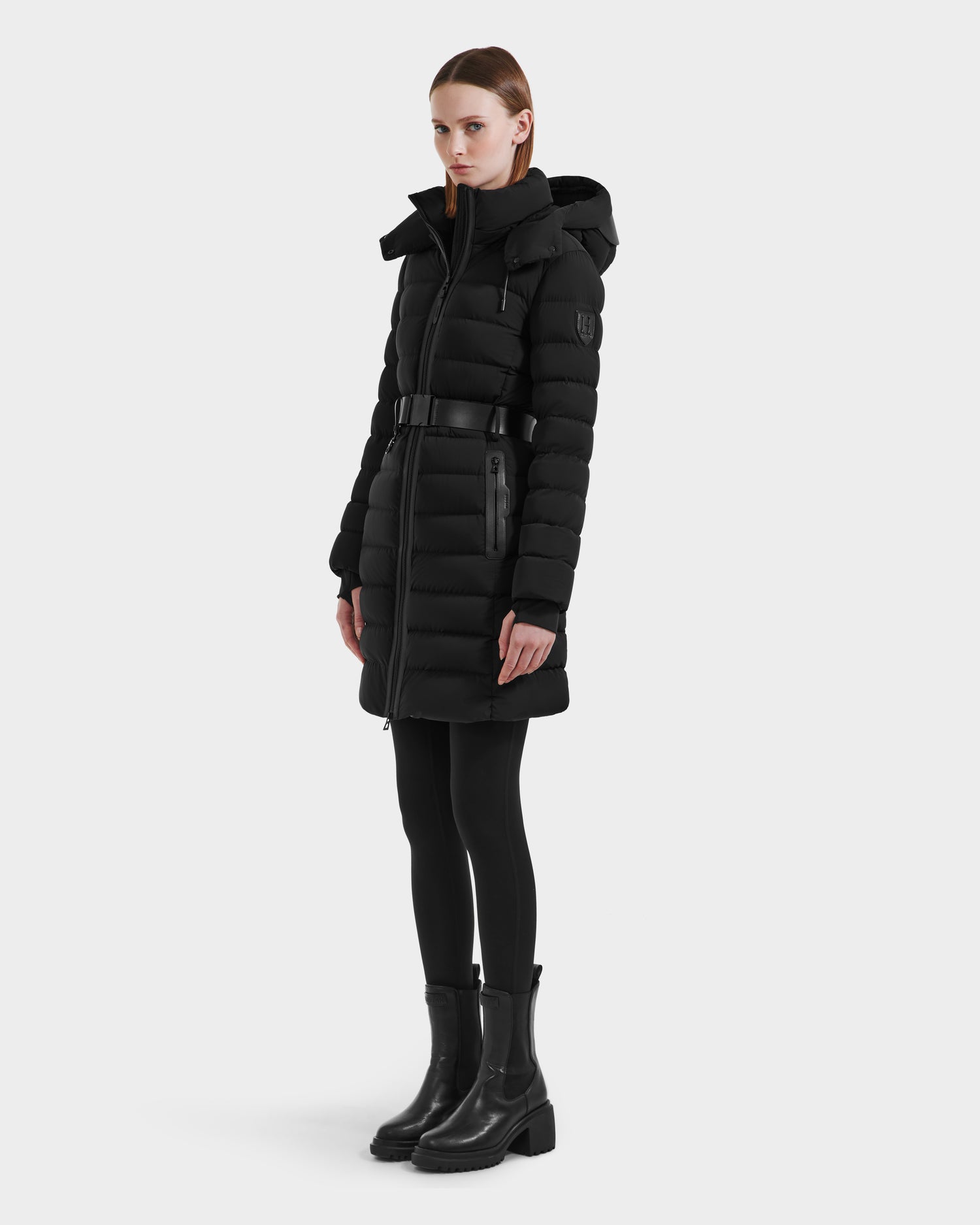 A person wears the Rudsak PELIN M HV - H125508 Black long, belted down jacket with a hood, stylish vegan leather accents, black leggings, and boots, standing slightly to the side against a plain light background.