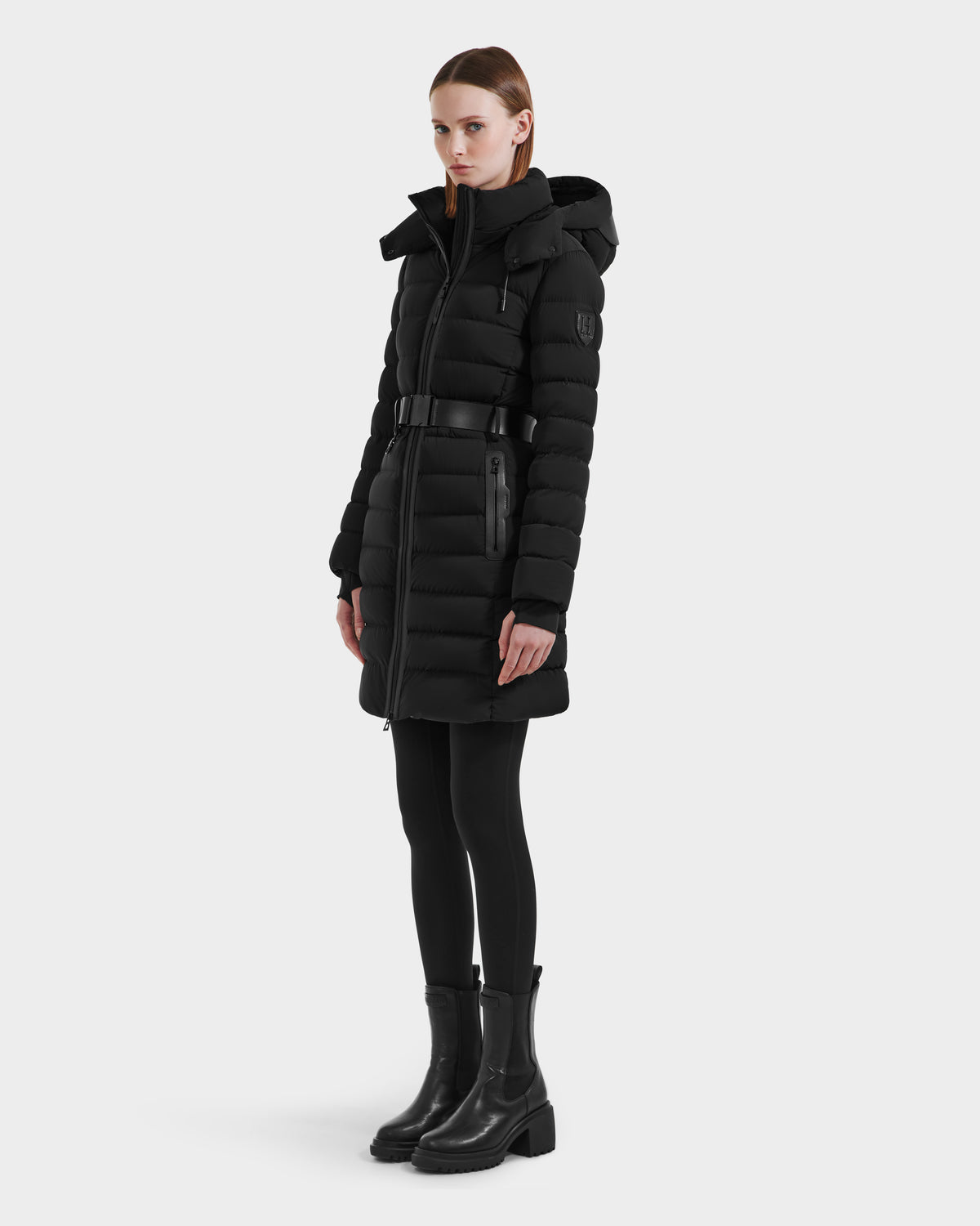 A person wears the Rudsak PELIN M HV - H125508 Black long, belted down jacket with a hood, stylish vegan leather accents, black leggings, and boots, standing slightly to the side against a plain light background.