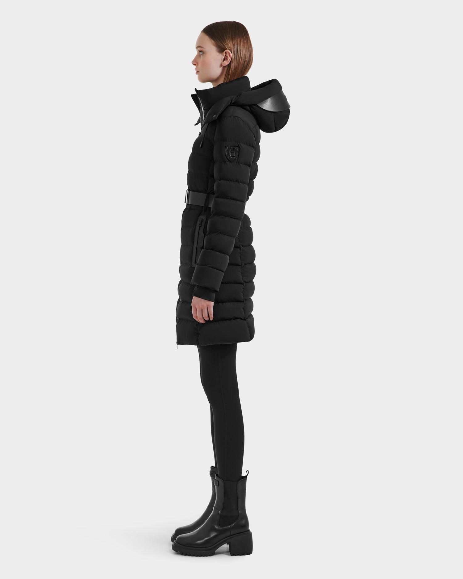 A woman stands in profile wearing Rudsak's PELIN M HV - H125508 Black belted vegan leather puffer coat with a hood, paired with black leggings and ankle boots, against a plain light gray background.