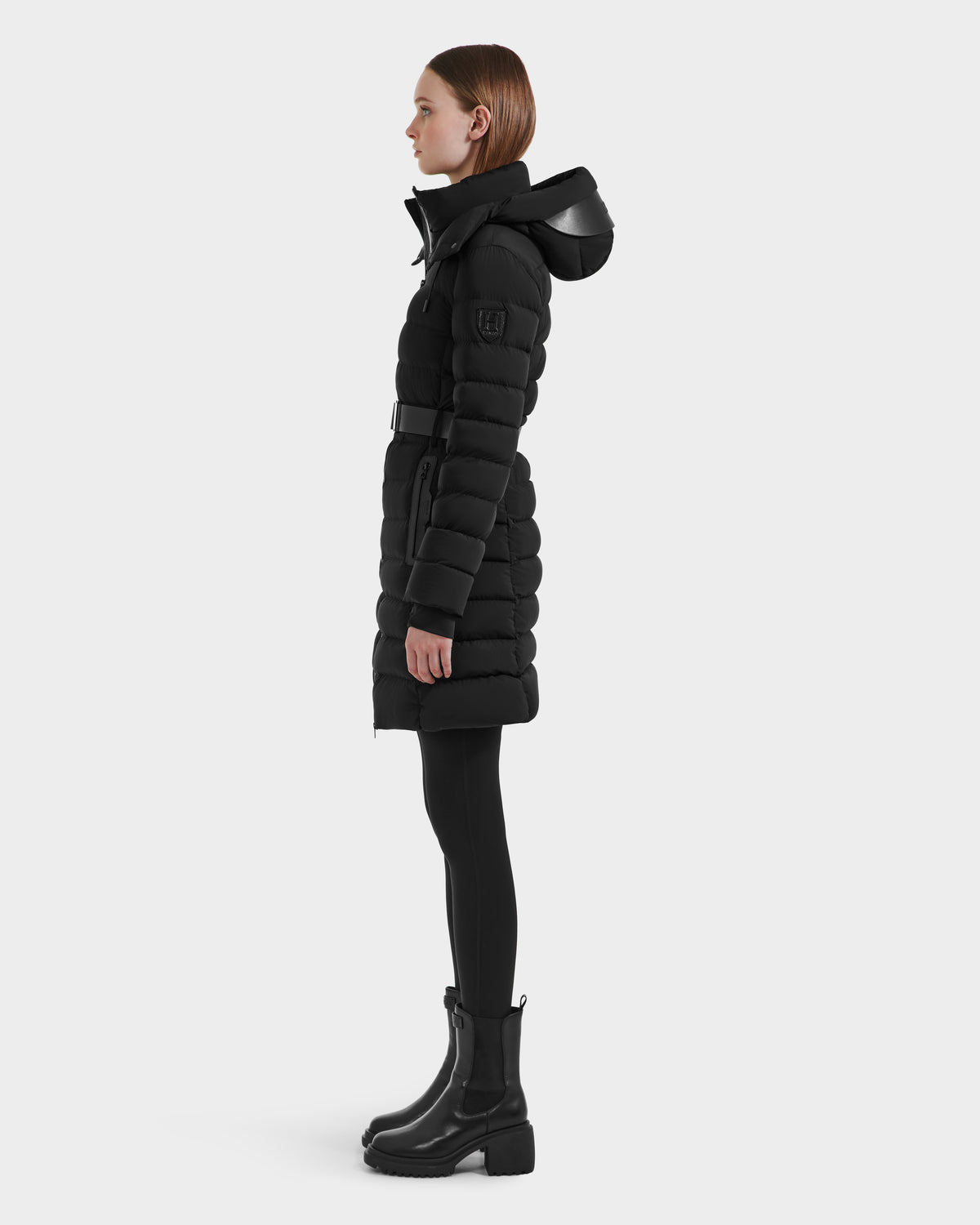 A woman stands in profile wearing Rudsak's PELIN M HV - H125508 Black belted vegan leather puffer coat with a hood, paired with black leggings and ankle boots, against a plain light gray background.