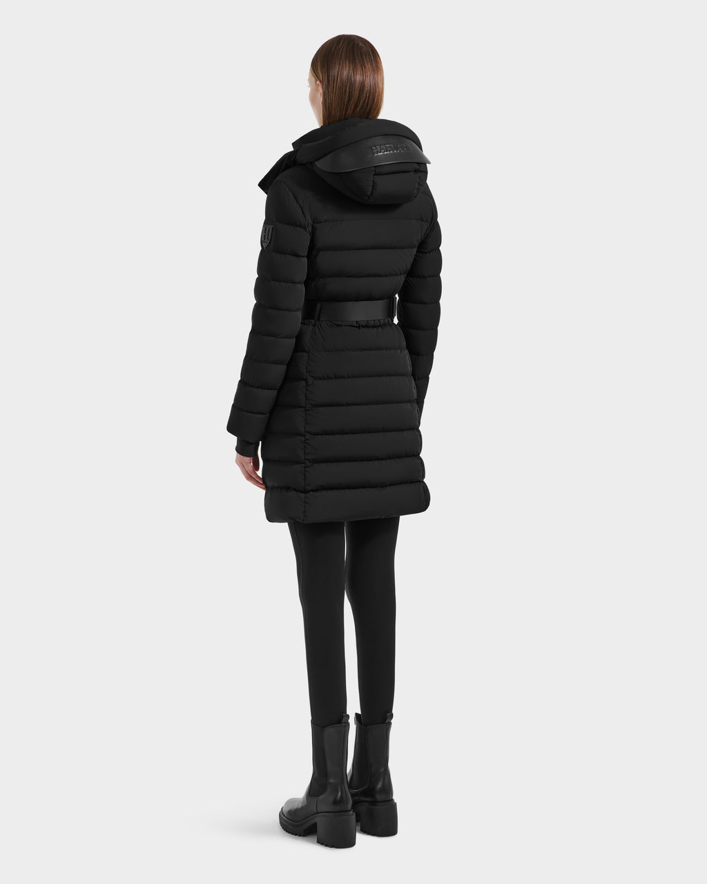 A person wearing the Rudsak PELIN M HV - H125508 Black vegan leather puffer coat with a hood, black leggings, and boots stands facing away from the camera against a plain light gray background.
