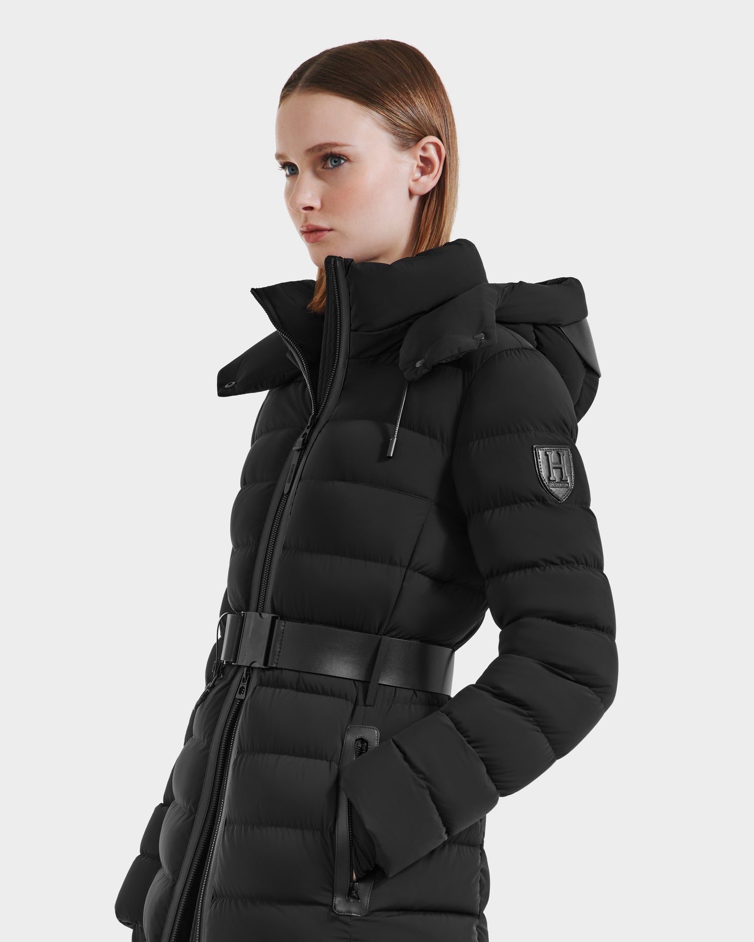 A woman with straight, shoulder-length brown hair wears the Rudsak PELIN M HV - H125508 Black belted puffer coat, featuring a high collar, hood, sleeve patch, and zippered pockets as she stands in profile against a plain background.