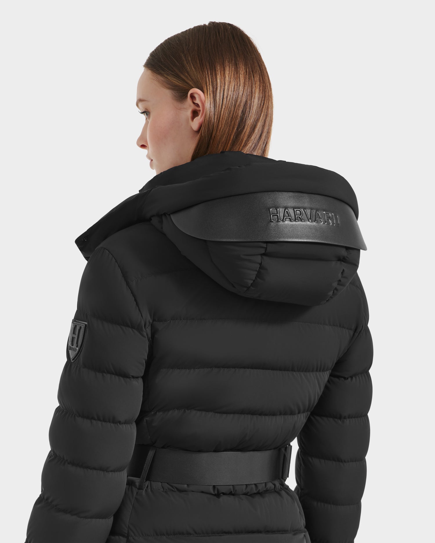 A woman with straight, light brown hair wears the Rudsak PELIN M HV - H125508 Black down jacket, featuring a hood with "HARVISH" in black, an upper arm patch, vegan leather accents, and a belted waist.