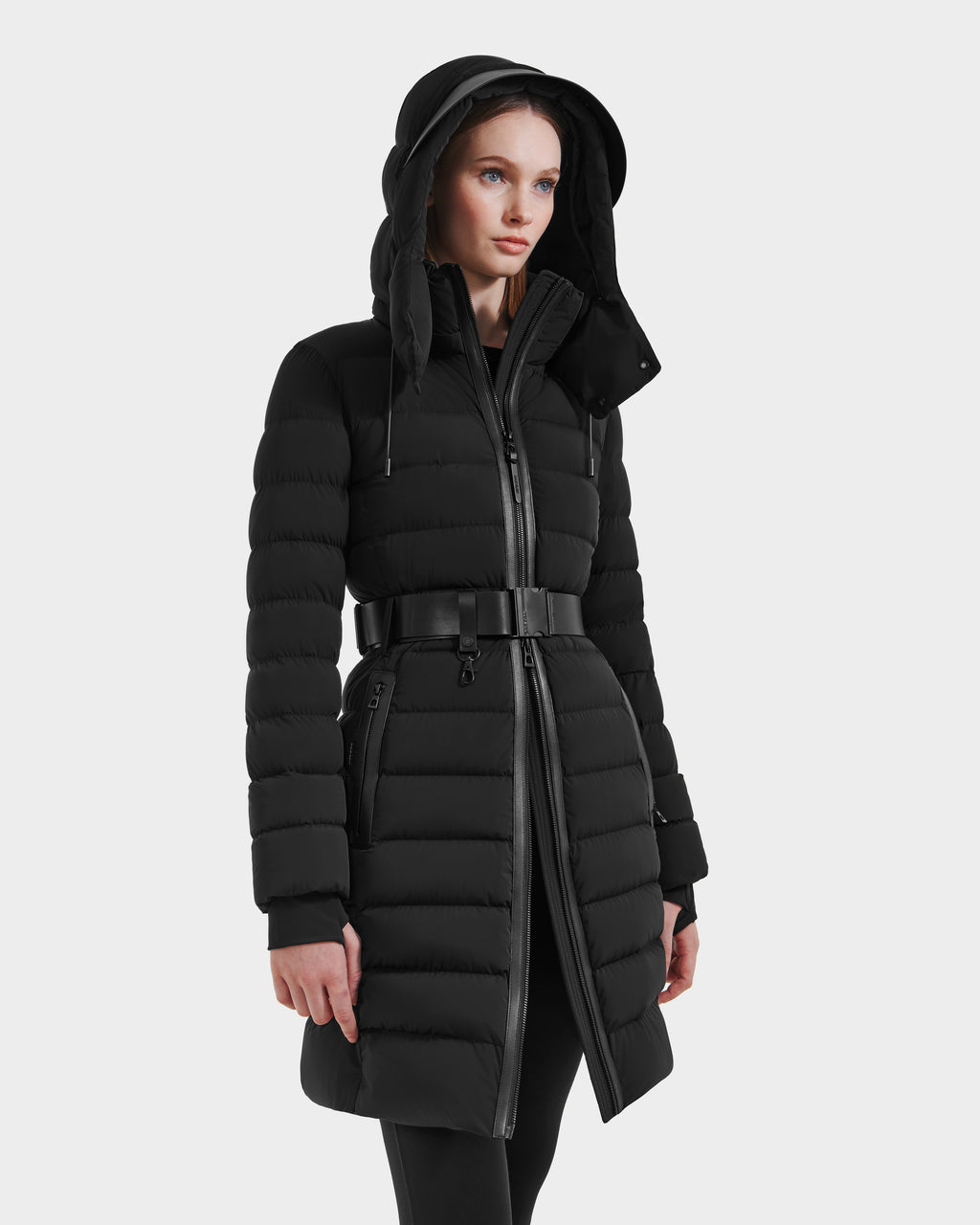 A woman wears the Rudsak PELIN M HV - H125508 Black: a long, black belted down jacket with a hood, front zipper, and horizontal quilted pattern, standing against a plain white background.